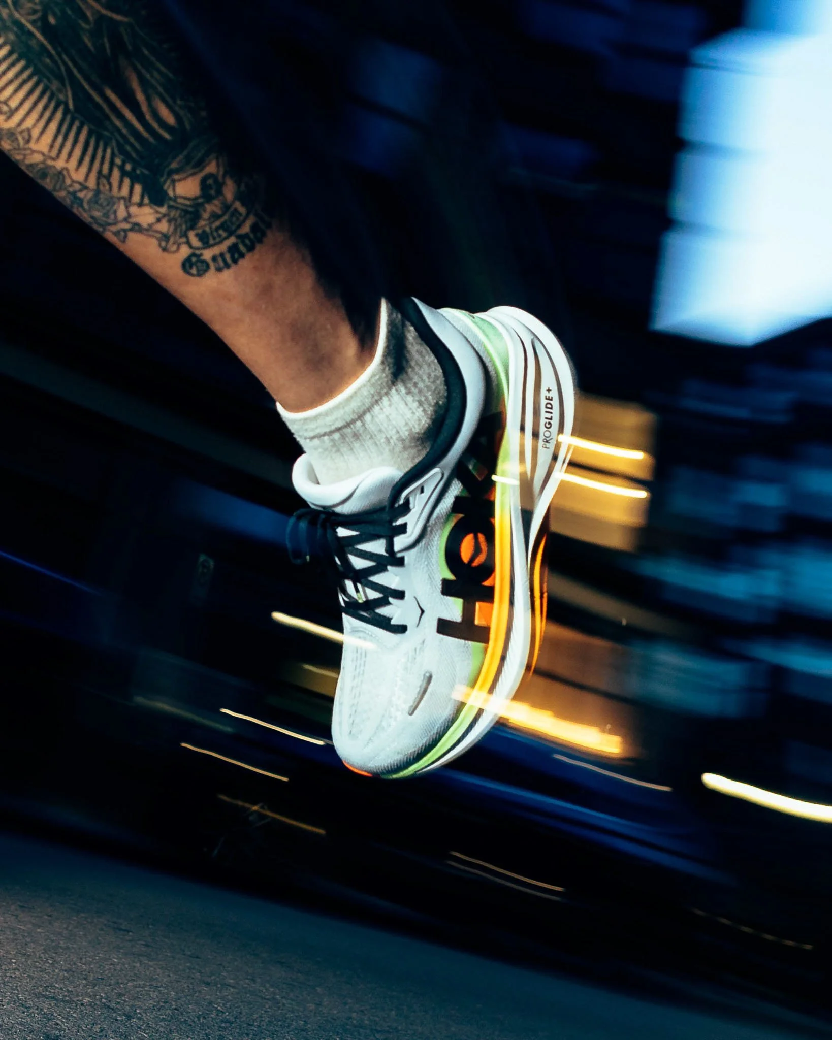 Blurry motion shot of a sports shoe on a dark road, with streaks of light indicating motion, and a tattooed leg visible.