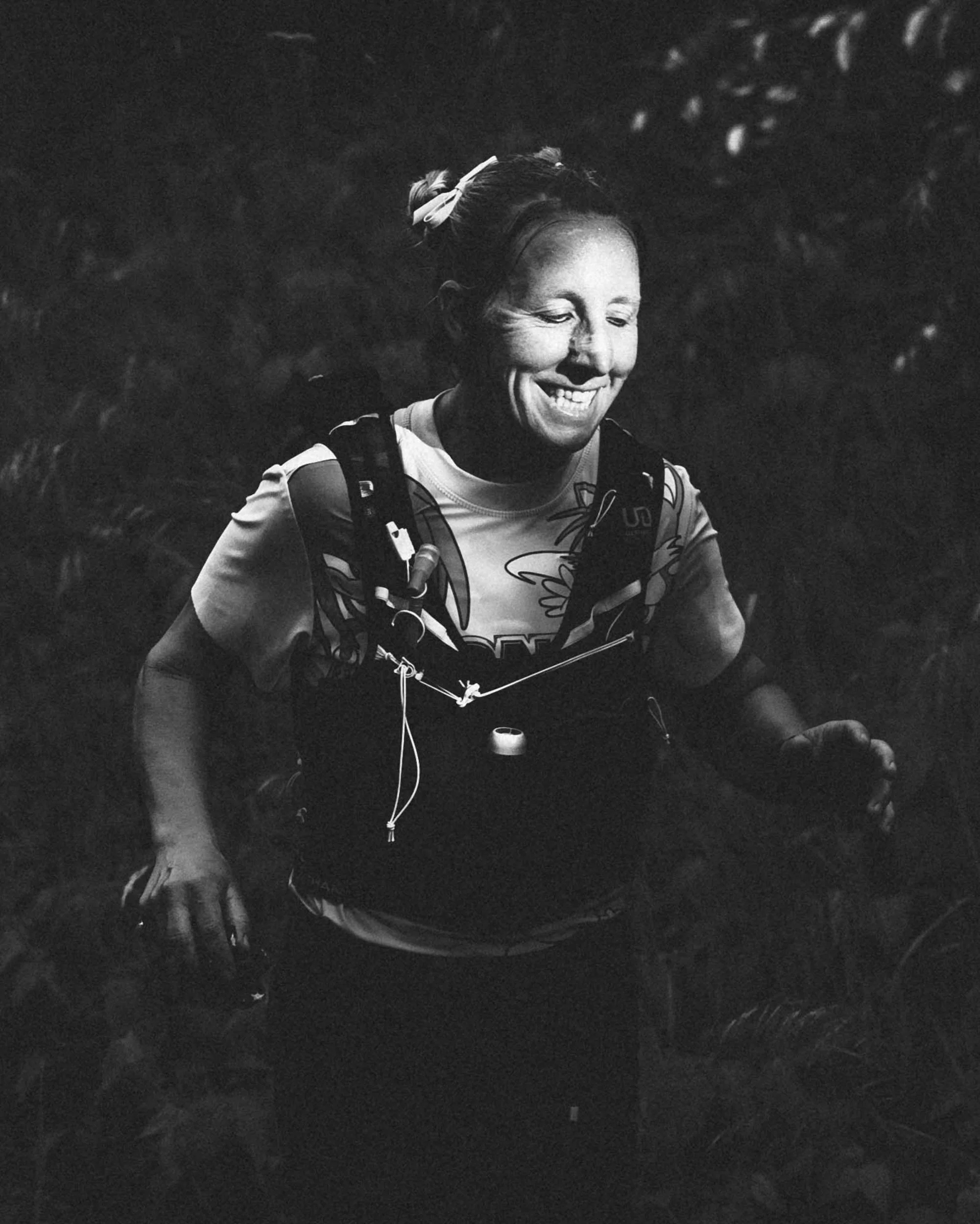 A woman smiling, wearing a sports shirt and a hydration vest, running on a trail outdoors with trees in the background, in black and white.