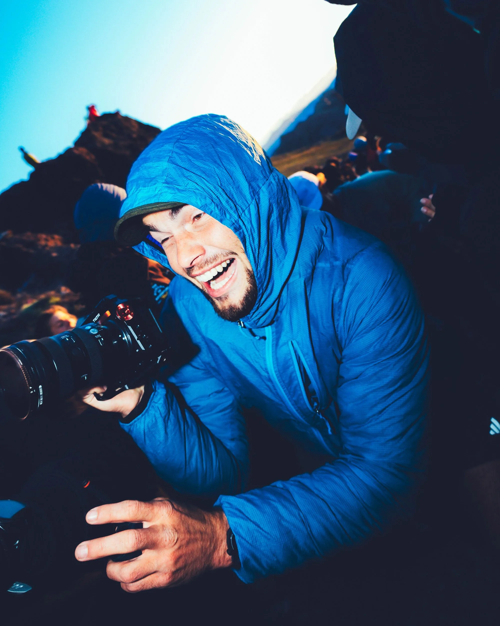 A person in a blue jacket and hood smiling as they hold a camera, surrounded by others outdoors near mountain terrain at sunset or sunrise.
