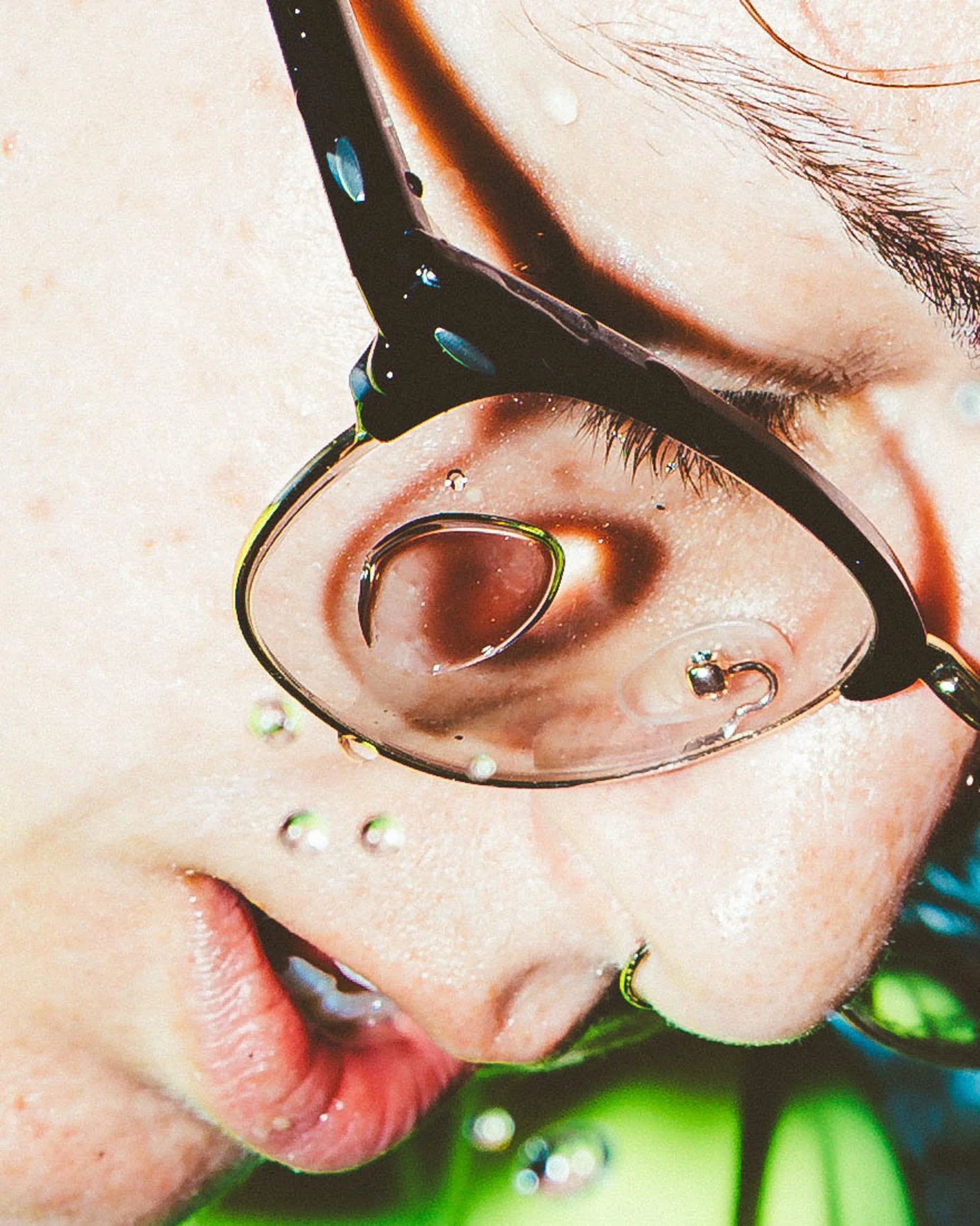 Close-up of a person with glasses and a piercing, with water droplets on their face.