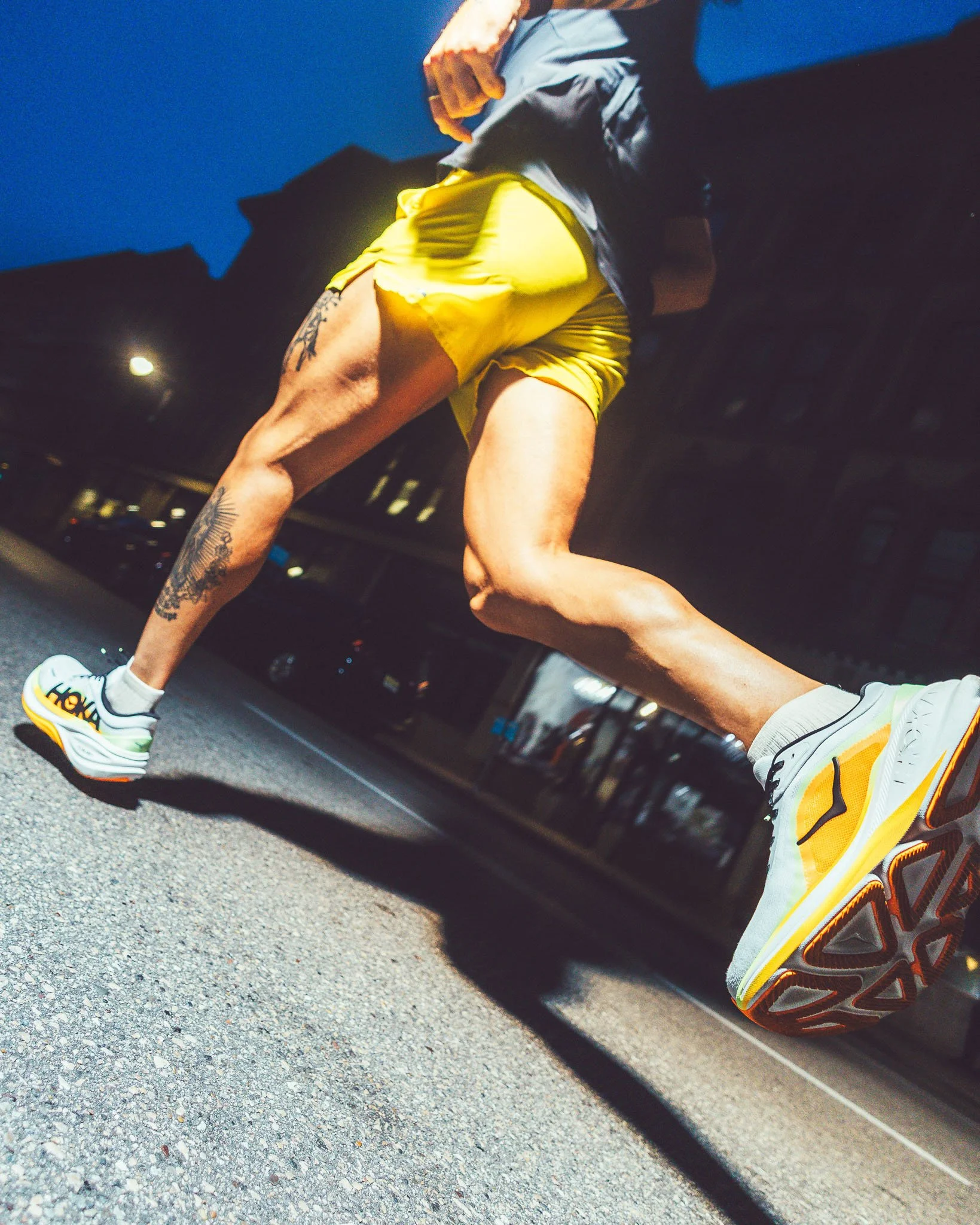 A man wearing a yellow athletic shirt and running shoes is running outdoors in the evening or early morning. The photo is taken from a low angle, focusing on his legs and shoes, with a dark building and streetlights in the background.