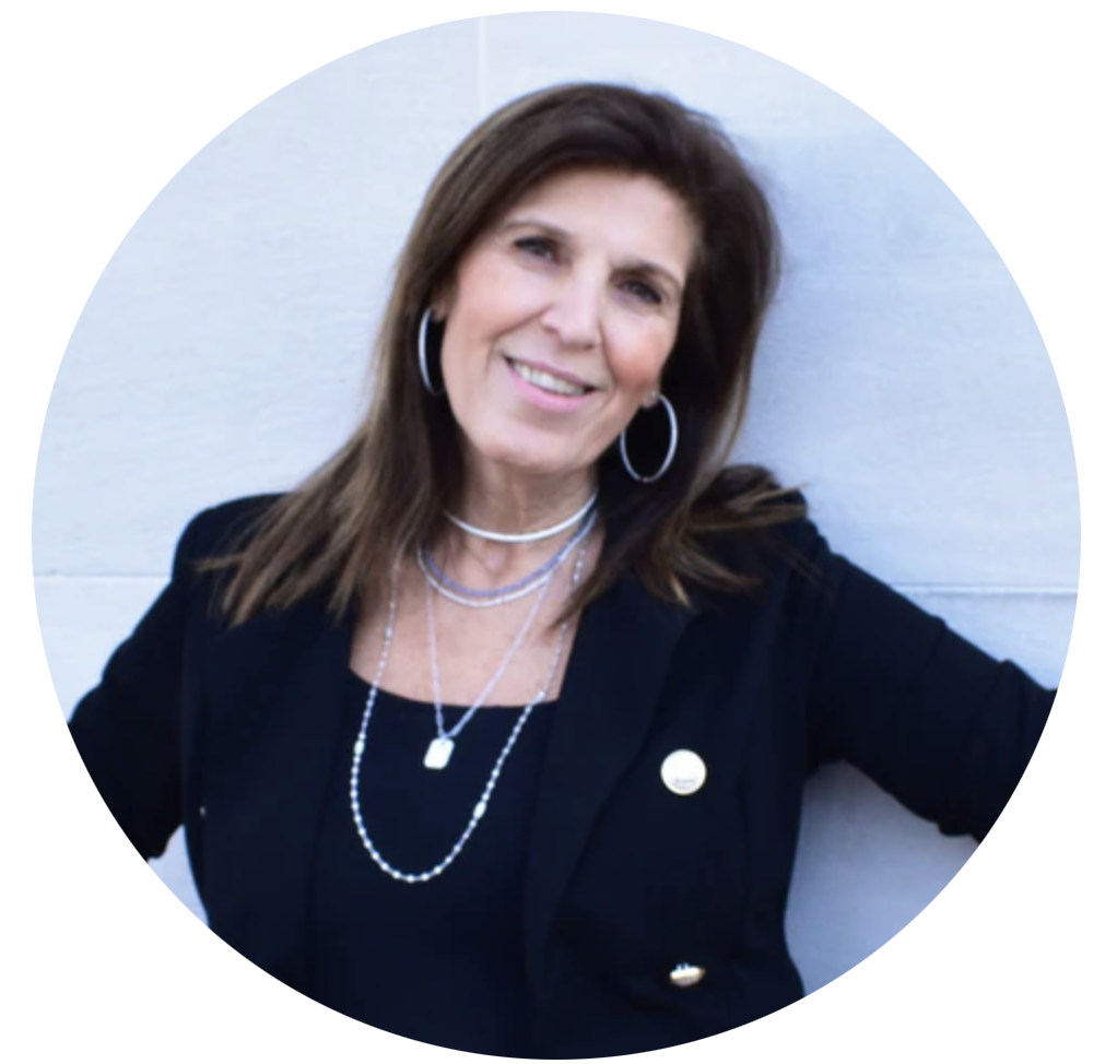 Smiling woman with long brown hair, wearing a black blazer and silver necklaces, leaning against a wall.