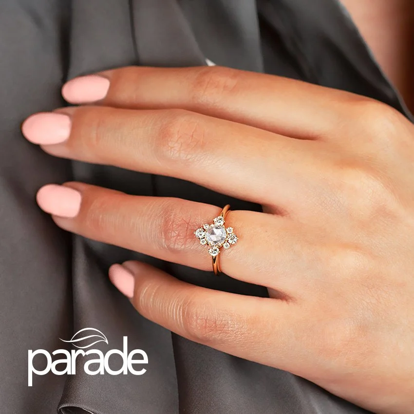 Close-up of a hand wearing a diamond engagement ring with a floral design on a gold band from Parade Jewelry, shown against a gray fabric background.