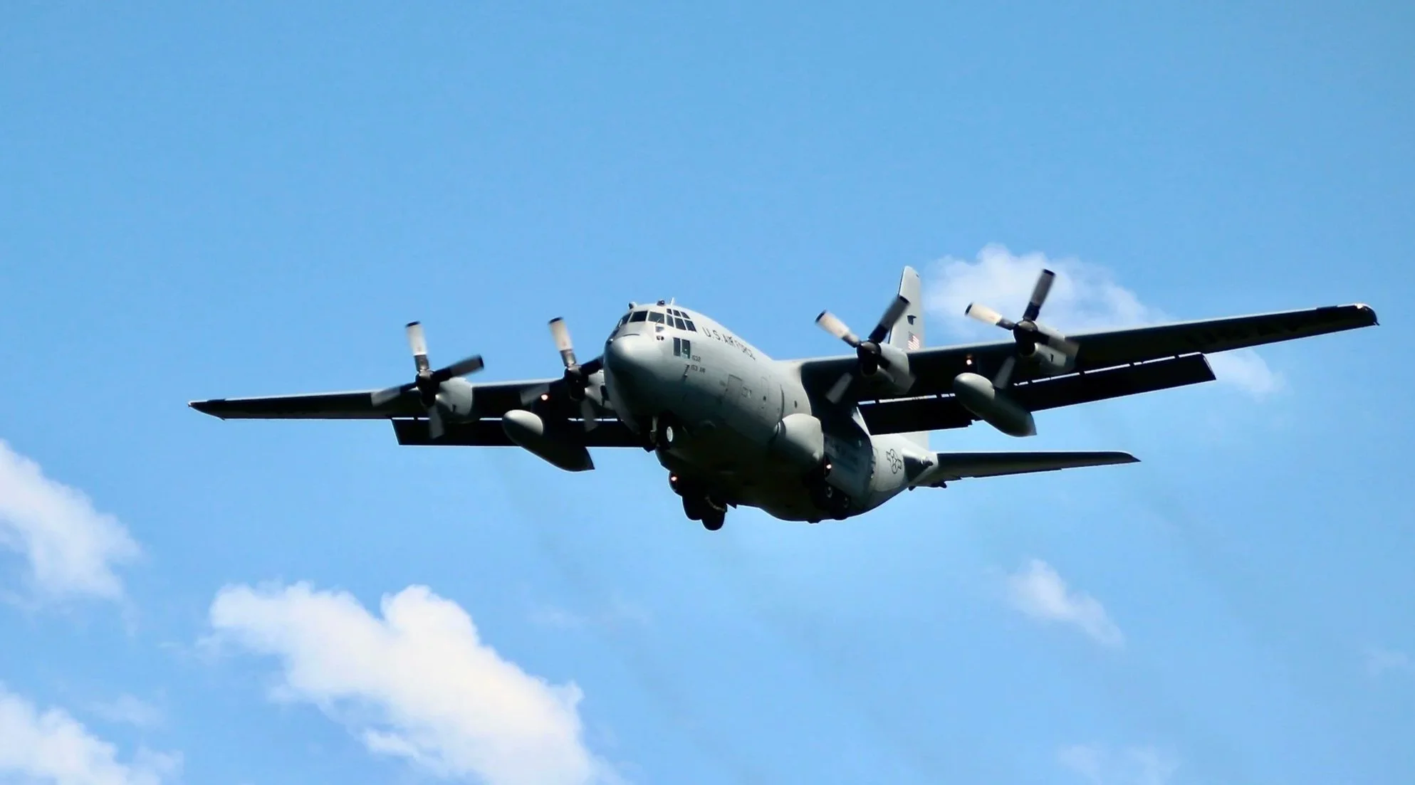 A large military transport aircraft flying in the blue sky with some clouds, propellers spinning.