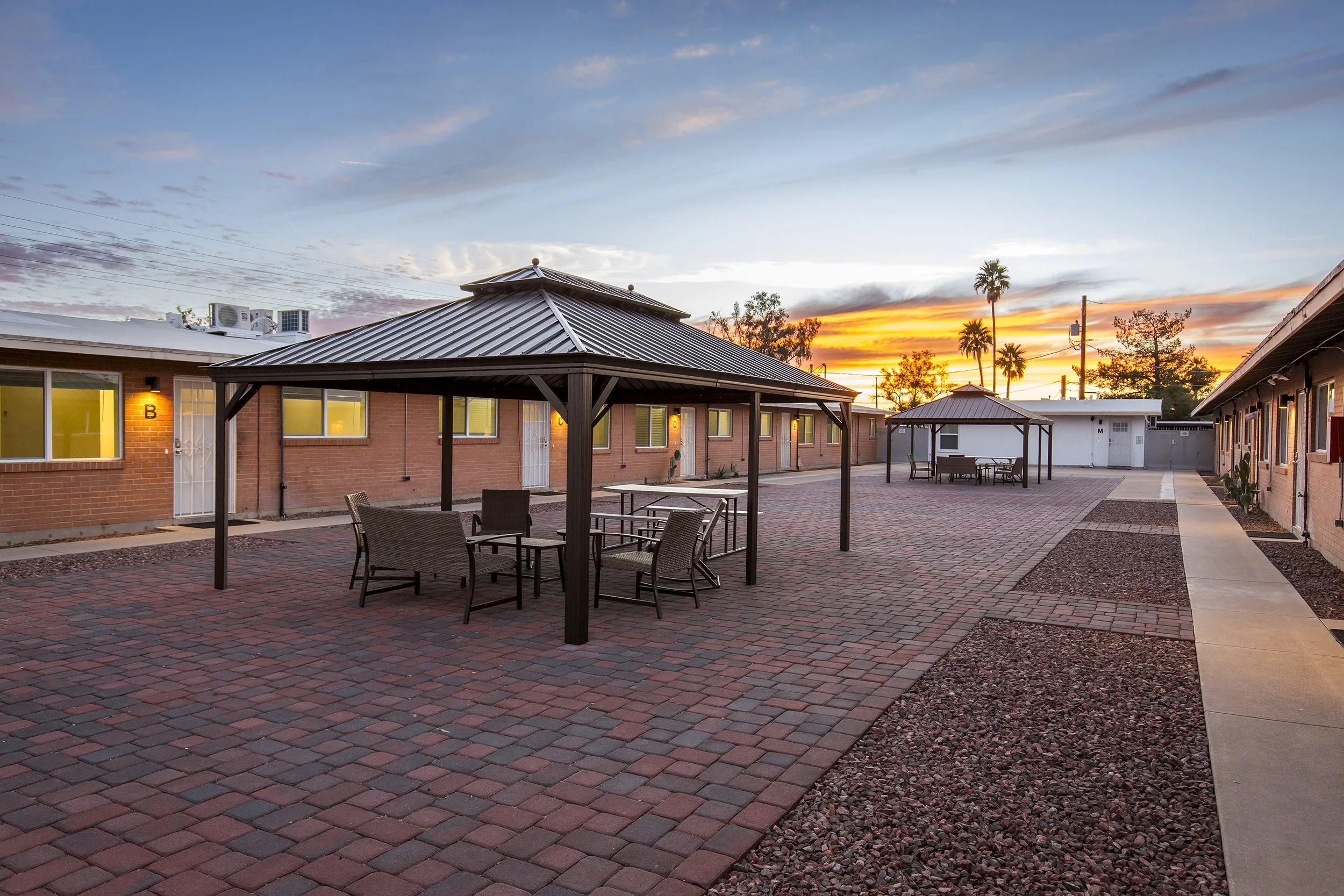 Brick courtyard with patio chairs and a ramada covering between brick apartment buildings at sunset.
