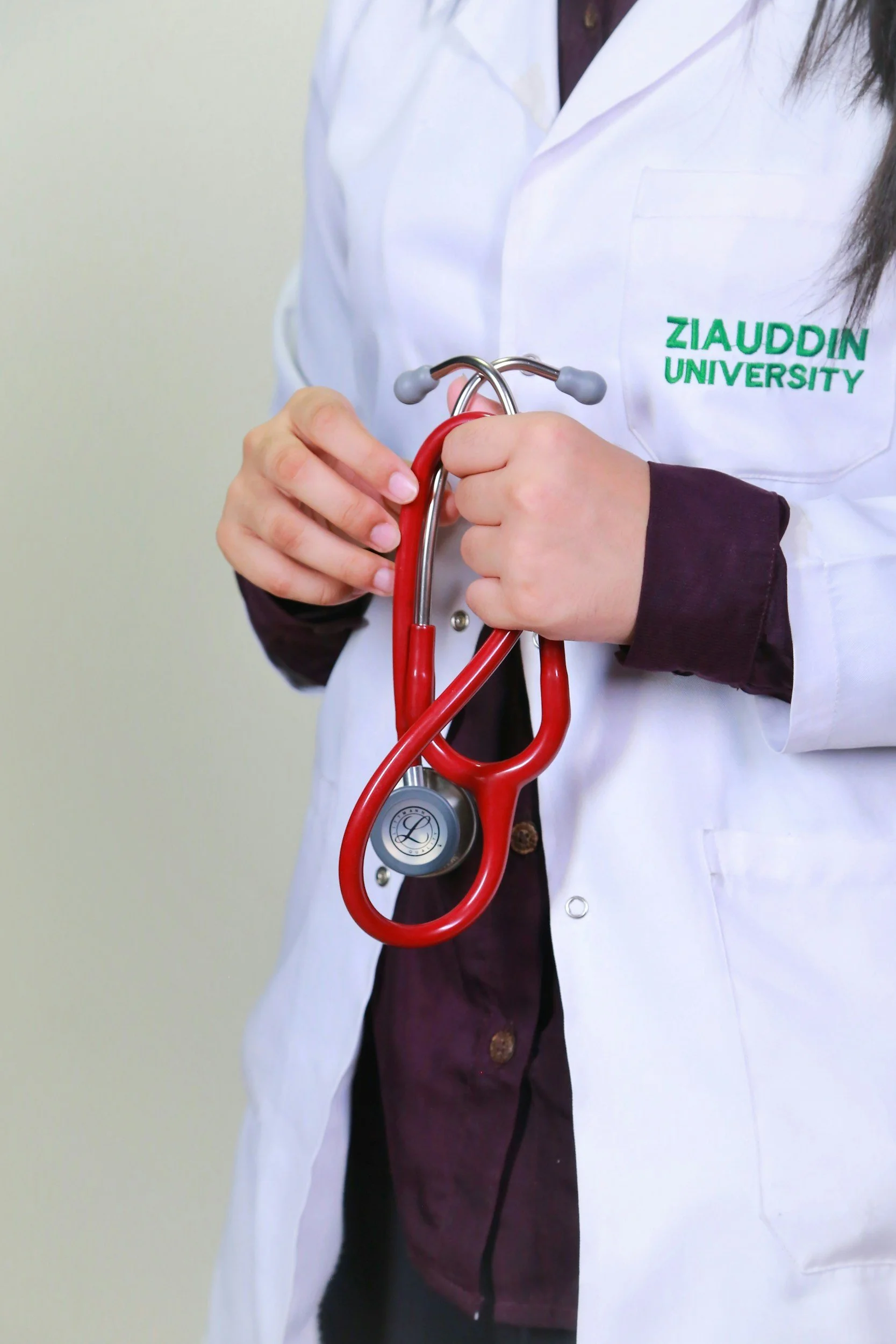 Person wearing a white lab coat with green text that reads 'ZIAUDDIN UNIVERSITY' holding a red stethoscope.