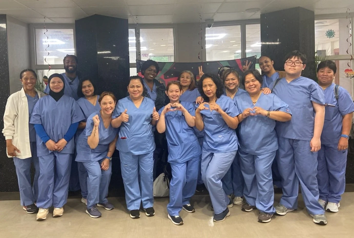 Group of healthcare workers in scrubs standing together indoors, some making heart shapes with their hands and smiling.