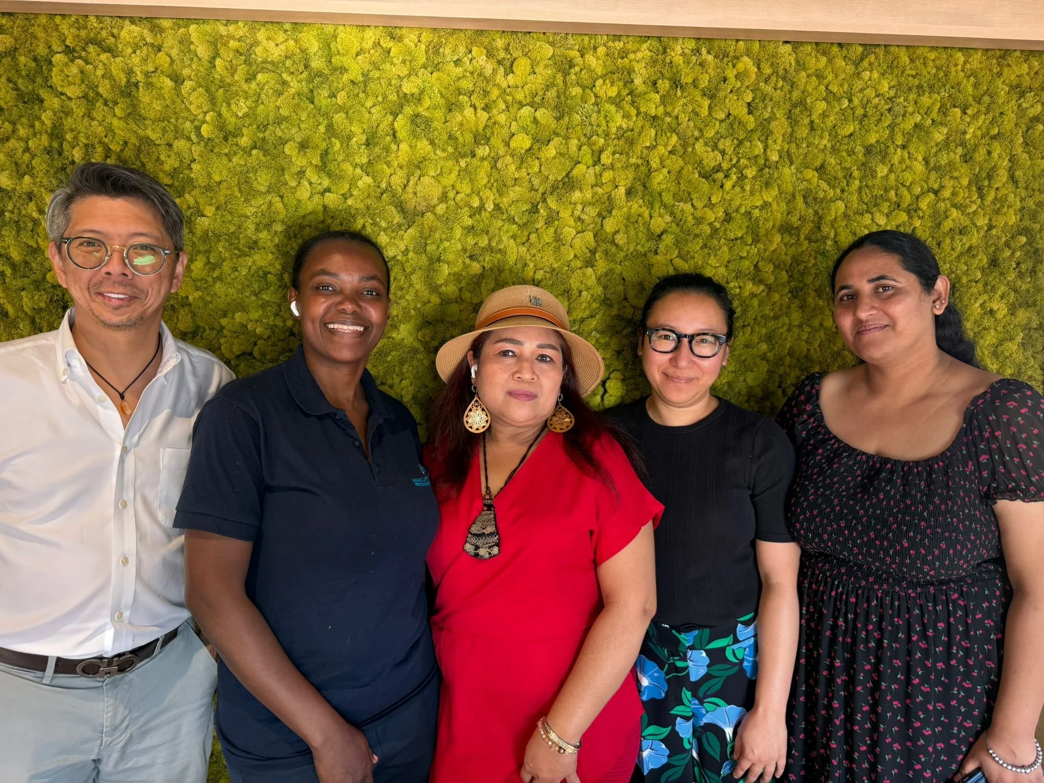 Group of five people standing in front of a green, textured wall, smiling and posing for the photo.