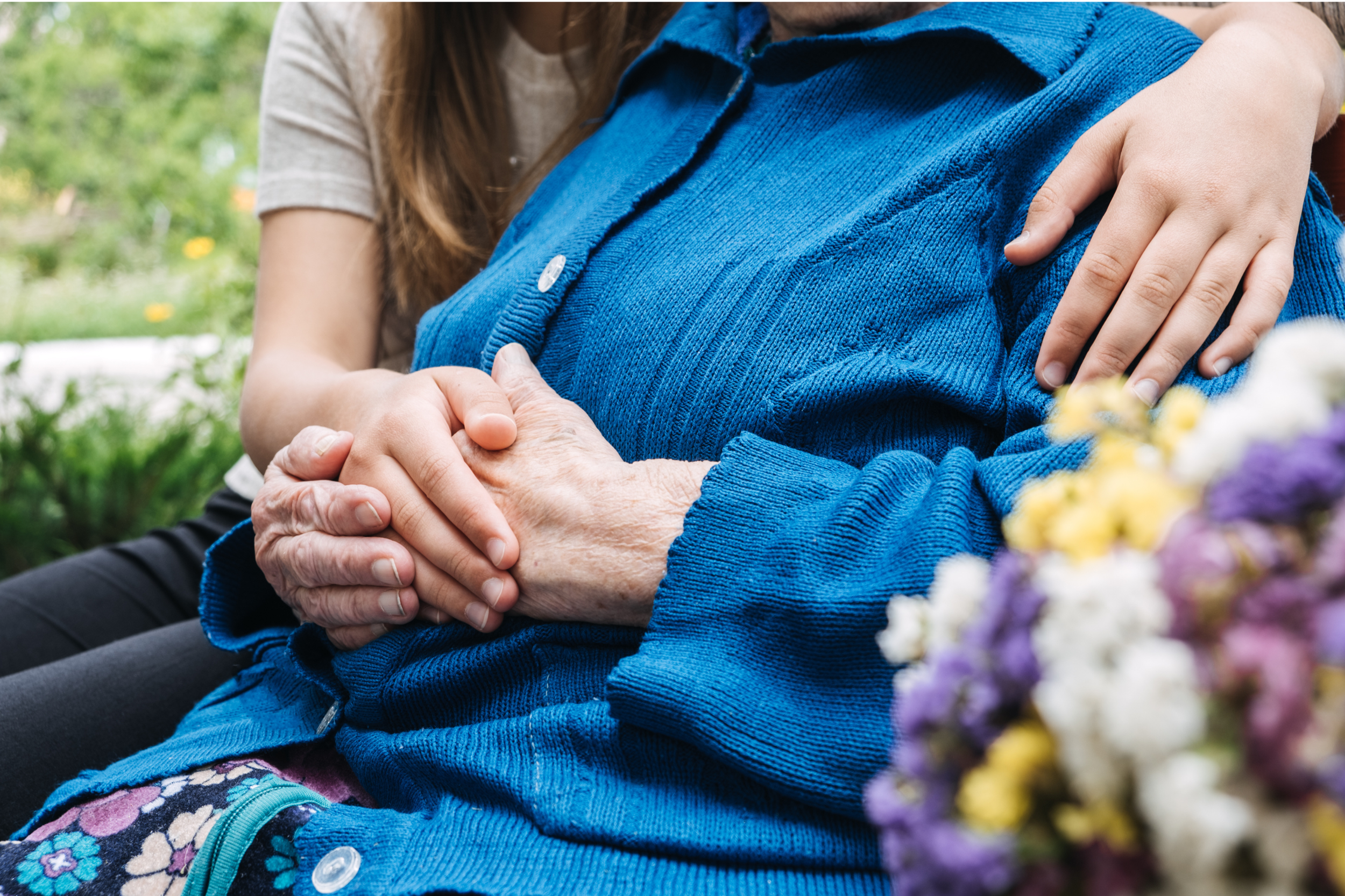 Two women sitting outdoors, one with an aged hand in the other's hand, with one woman's arm around the other's shoulder, surrounded by greenery and a bouquet of flowers.