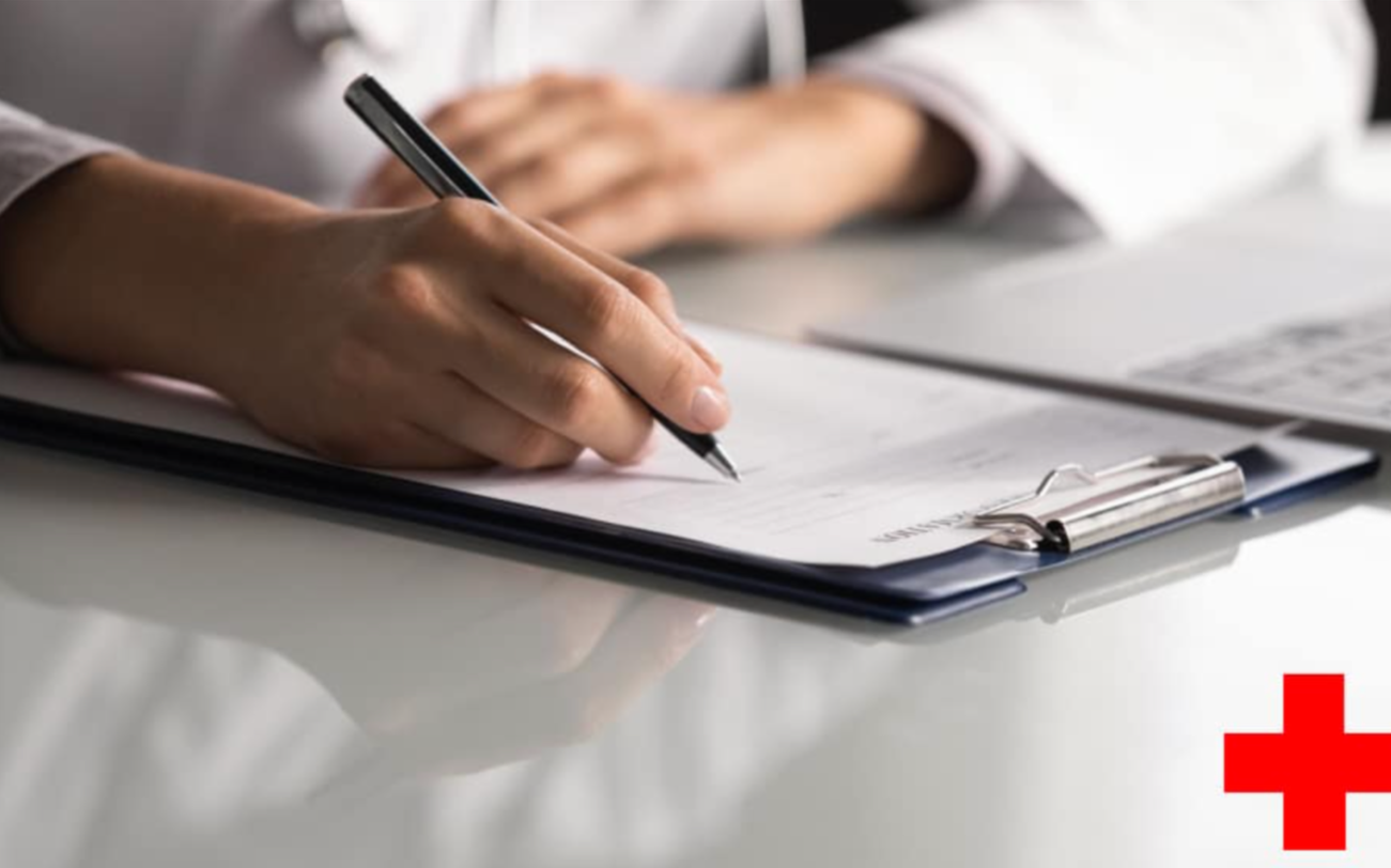 Person writing on a clipboard with medical forms, resting on a white desk, with a red cross symbol in the bottom right corner.