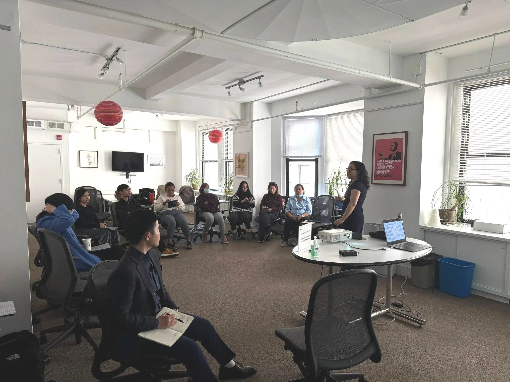A woman standing and speaking to a seated group of diverse people in a brightly lit office conference room with large windows, blinds, and wall art.