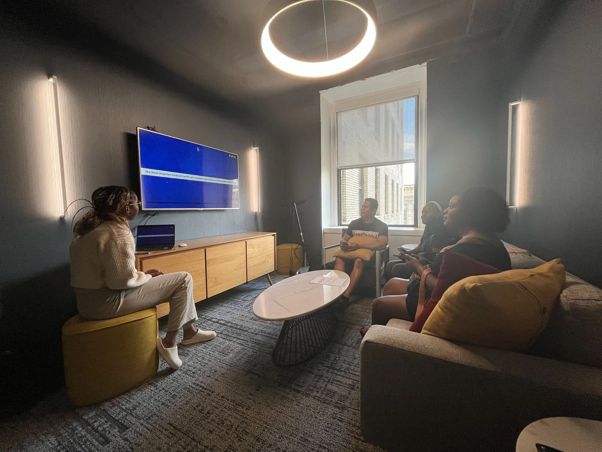 A group of four people sitting on a gray sofa and a yellow ottoman in a living room, watching a woman giving a presentation or lecture that is projected on a large blue screen on the wall.