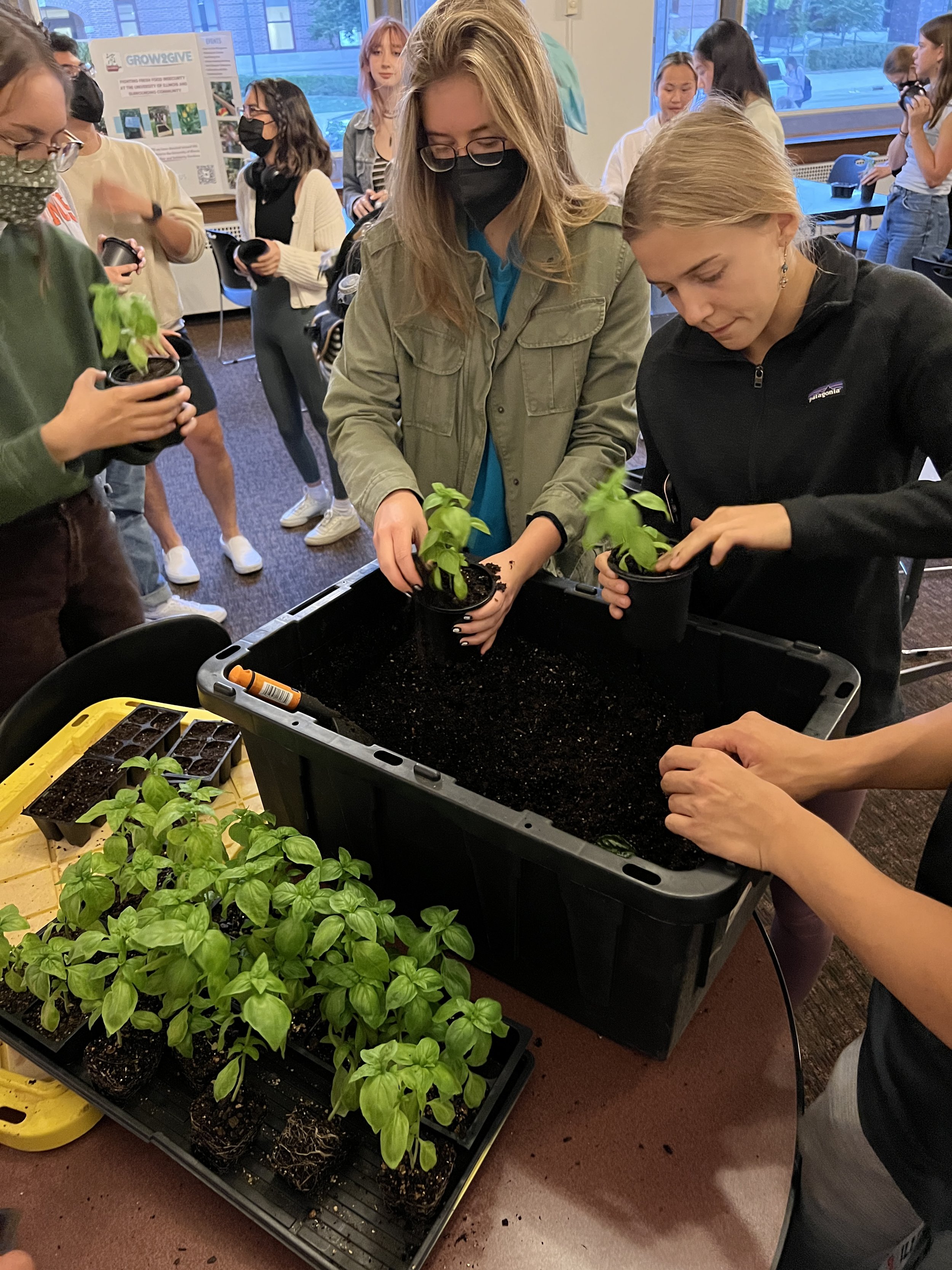 A Grow2Give event with people planting seedlings in small pots at a table indoors, with a group of onlookers in the background.