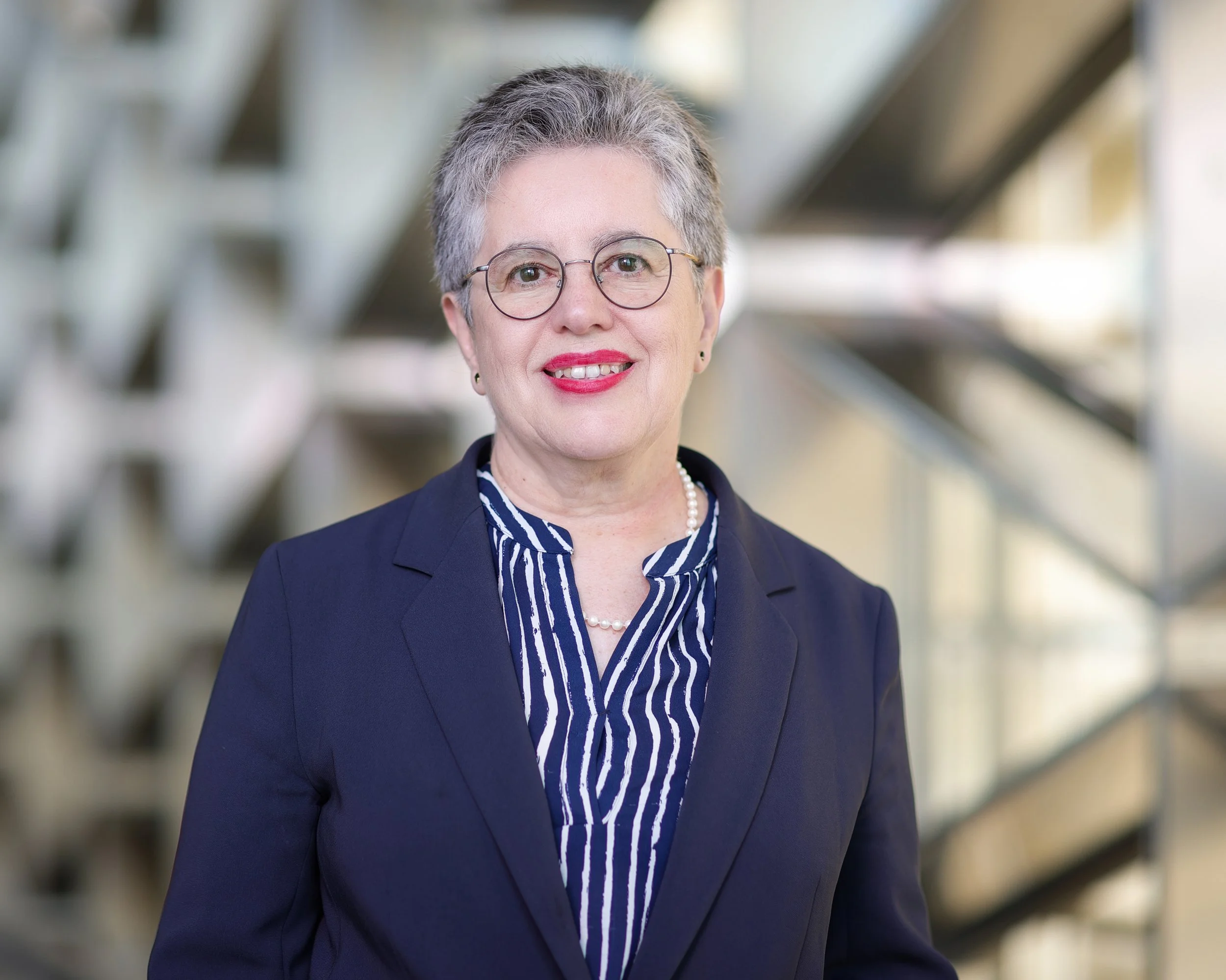 A professional woman with short gray hair, glasses, and red lipstick wearing a navy blazer, pearl necklace, and striped blouse, standing indoors with metal stairs in the background.