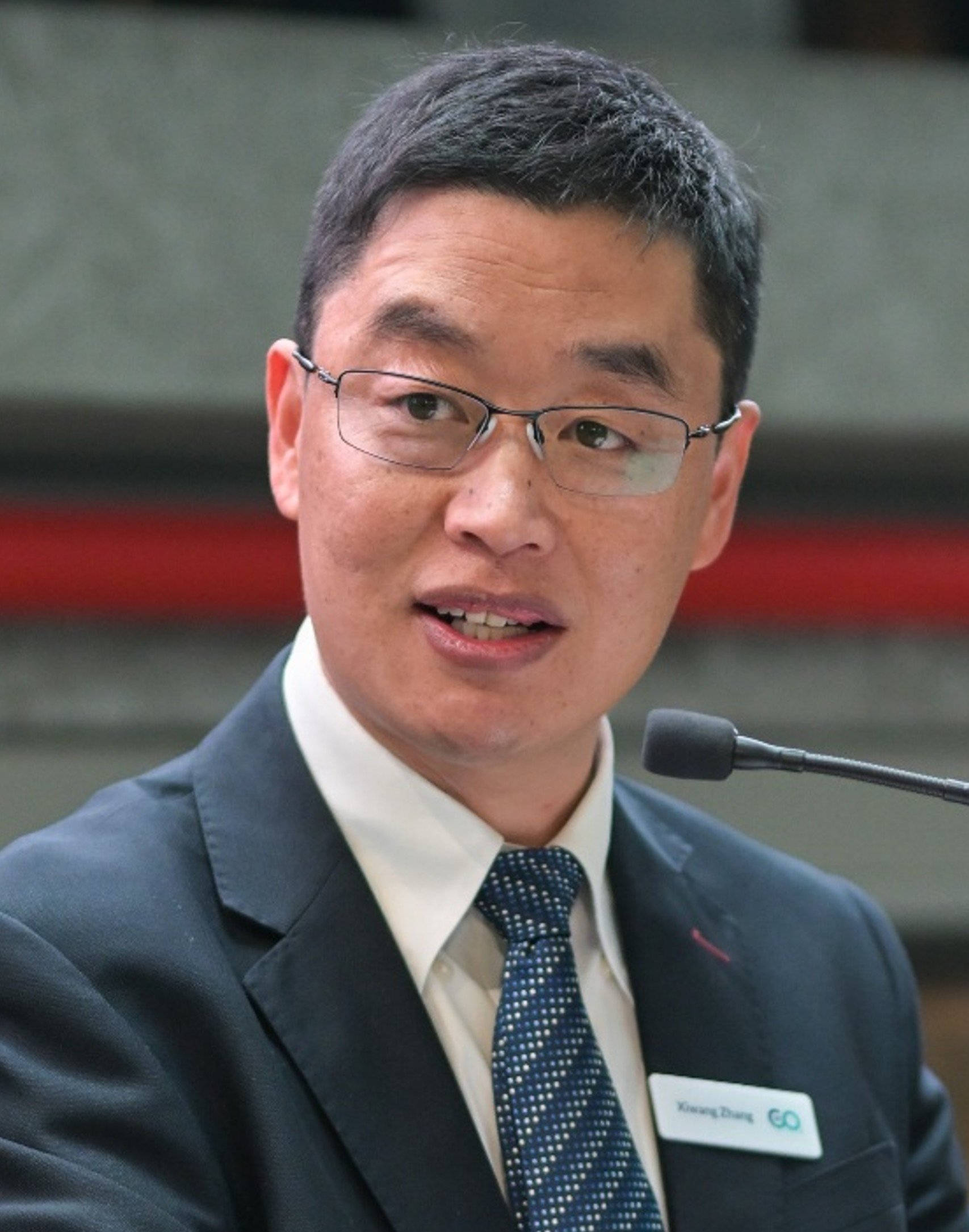 Man speaking into a microphone wearing glasses, a suit, and a name tag.