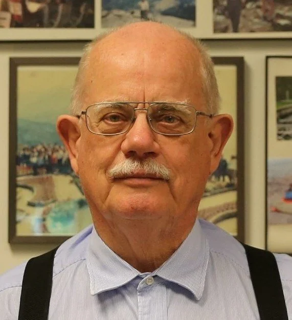 An elderly man with glasses and a mustache, wearing a light blue collared shirt and black suspenders, standing in front of a wall with framed photographs.
