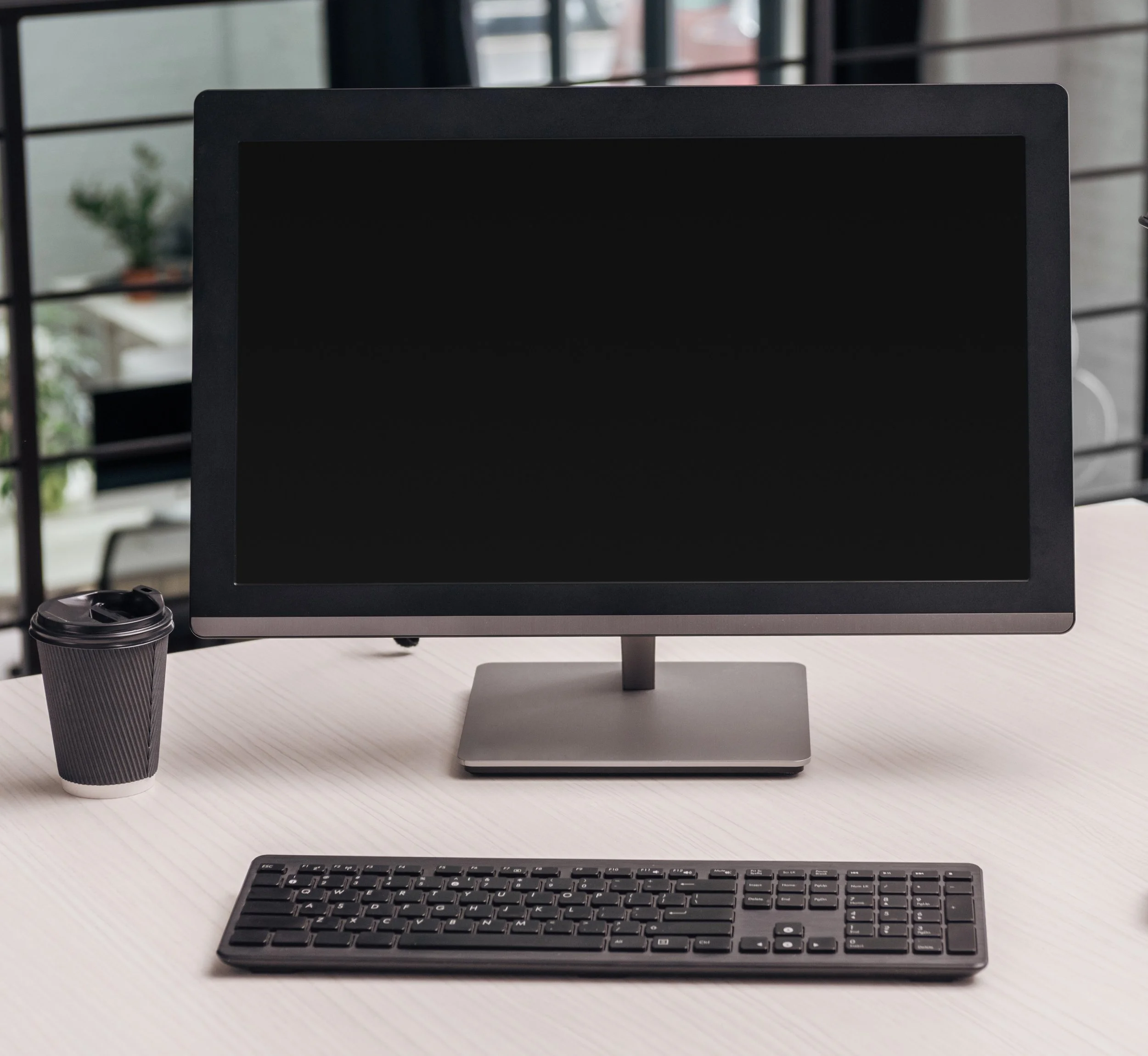 Computer monitor, keyboard, and a paper coffee cup on a white desk in an office setting.