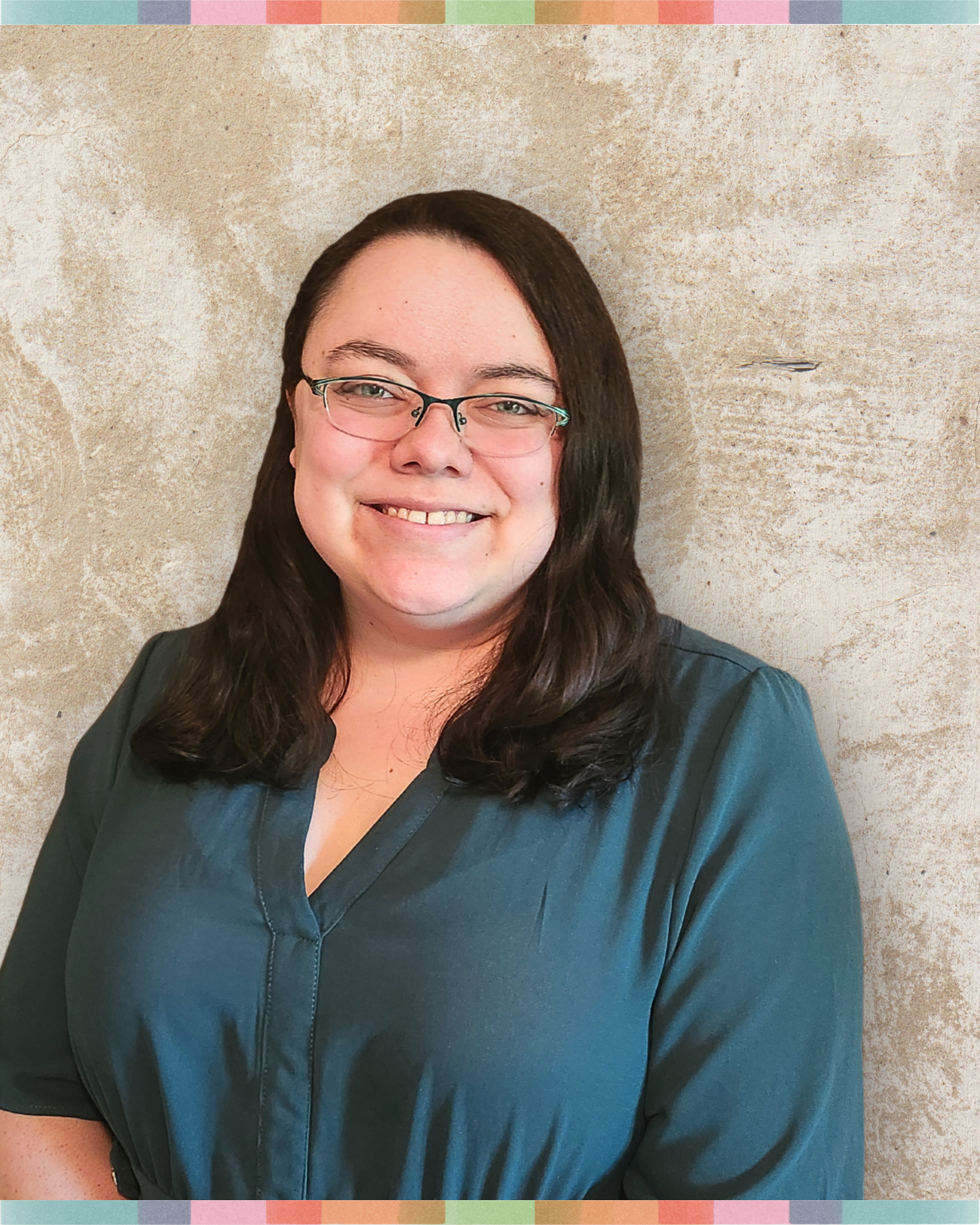A woman with long brown hair, glasses, and light skin, smiling in front of a textured beige wall, wearing a dark teal top.