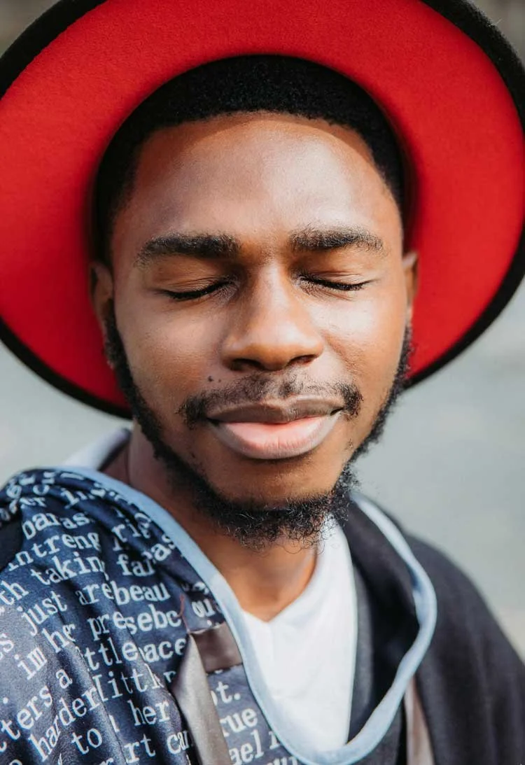 A young man with closed eyes wearing a red hat with a wide brim and a blue and white patterned jacket.