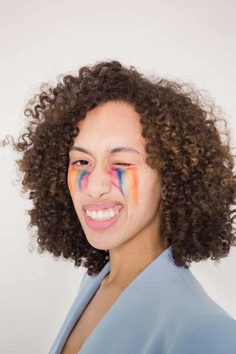 Woman with curly hair winking and smiling, face painted with rainbow-colored streaks.