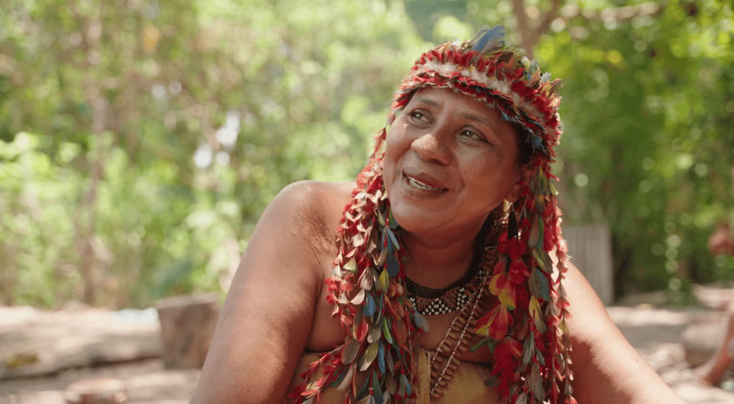 Suluene, smiling and wearing ceremonial garb, speaks to the camera while looking at the environment around her
