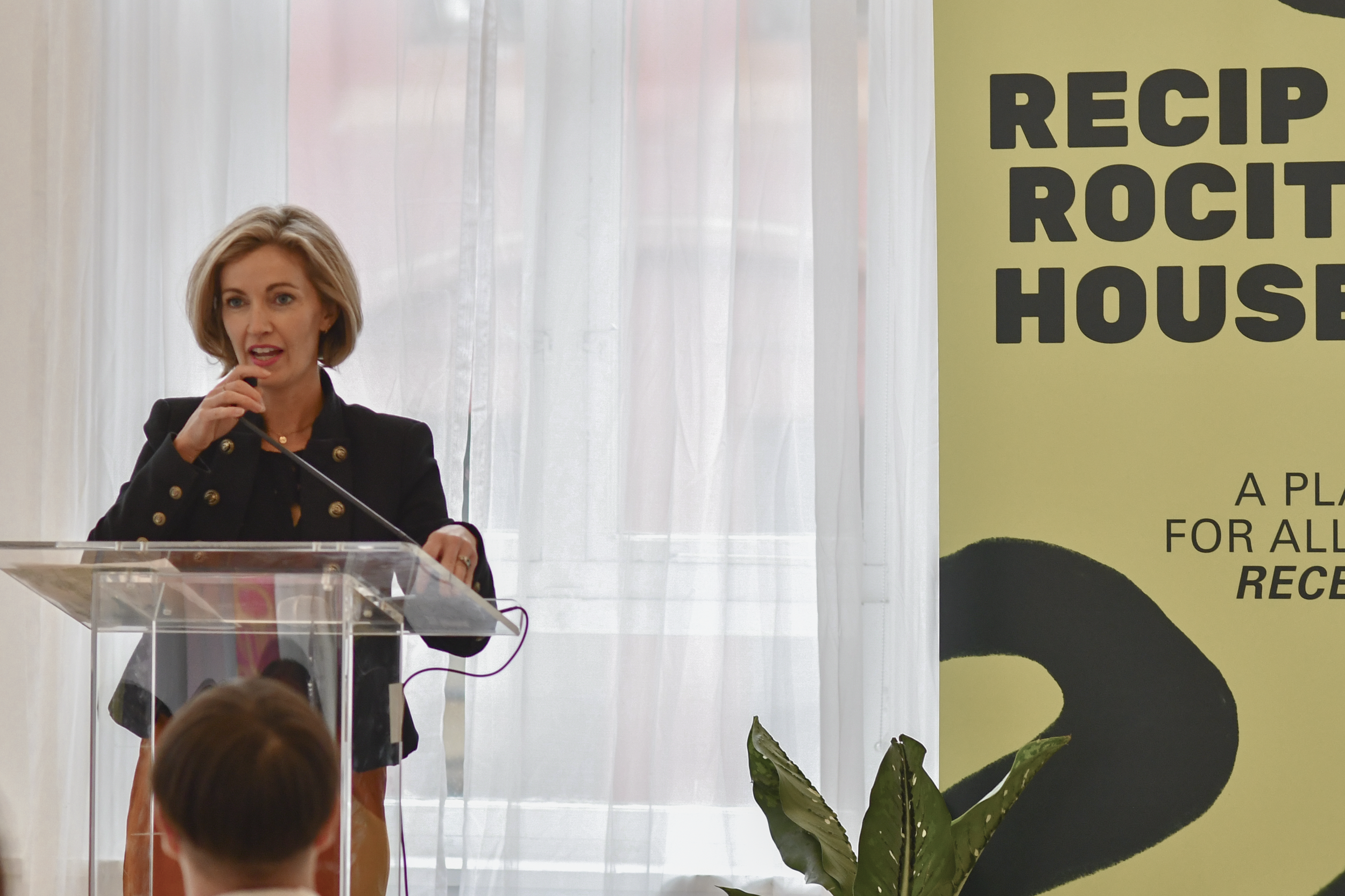Ashley Emerson speaks on a clear podium. She is wearing a black blazer and orange skirt and is holding a microphone. Beside her, a large banner says 'RECIPROCITY HOUSE'