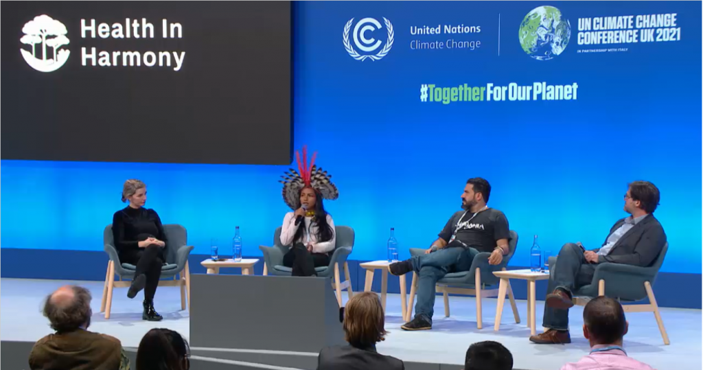 Juma Xipaia speaks at COP26 stage, with Ashley Emerson, Marcelo Salazar, and Jonathan Jennings, on a Health In Harmony stage