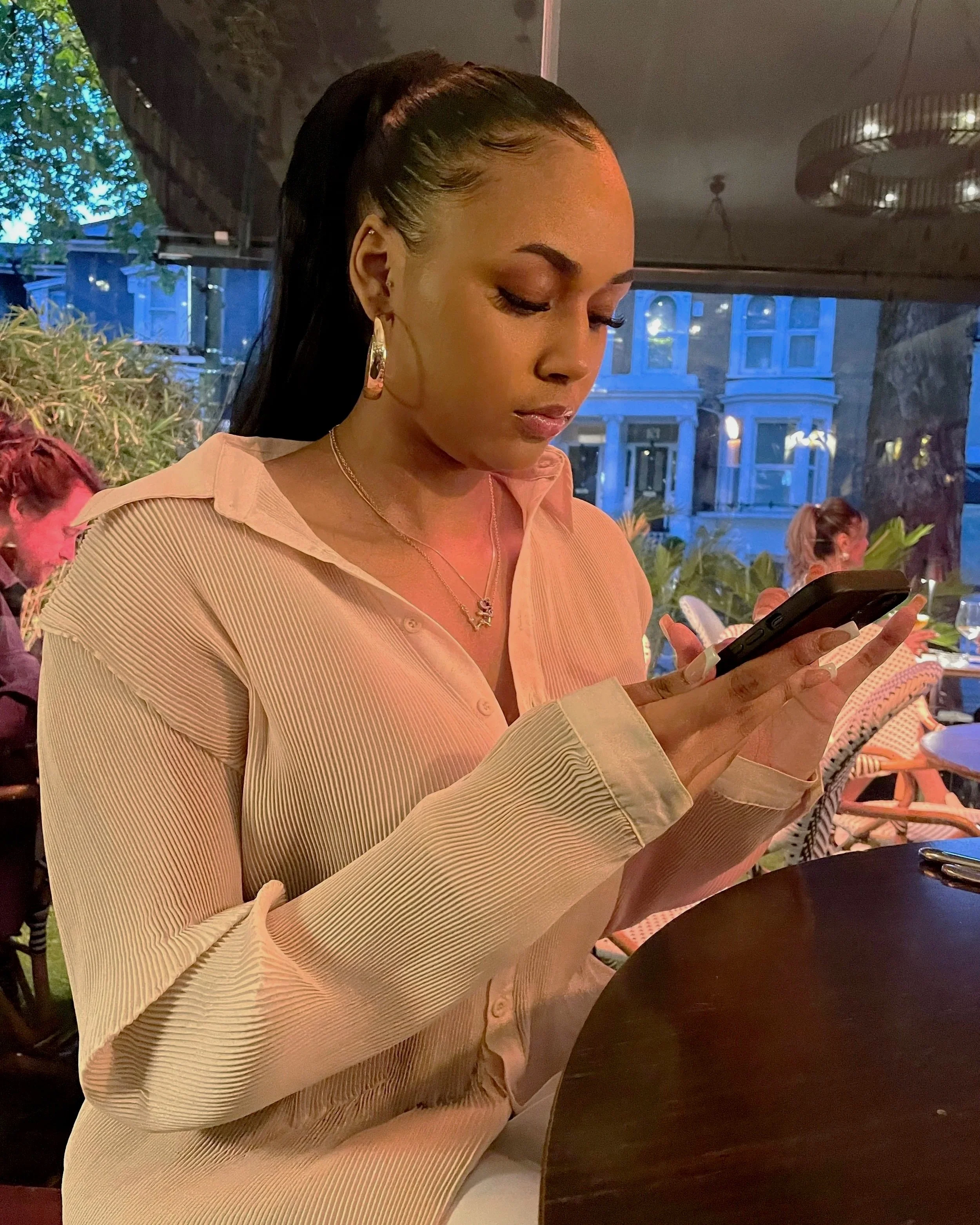 Image of young woman, scrolling on her phone whilst seated at a restaurant table, large window behind her with others dining.
