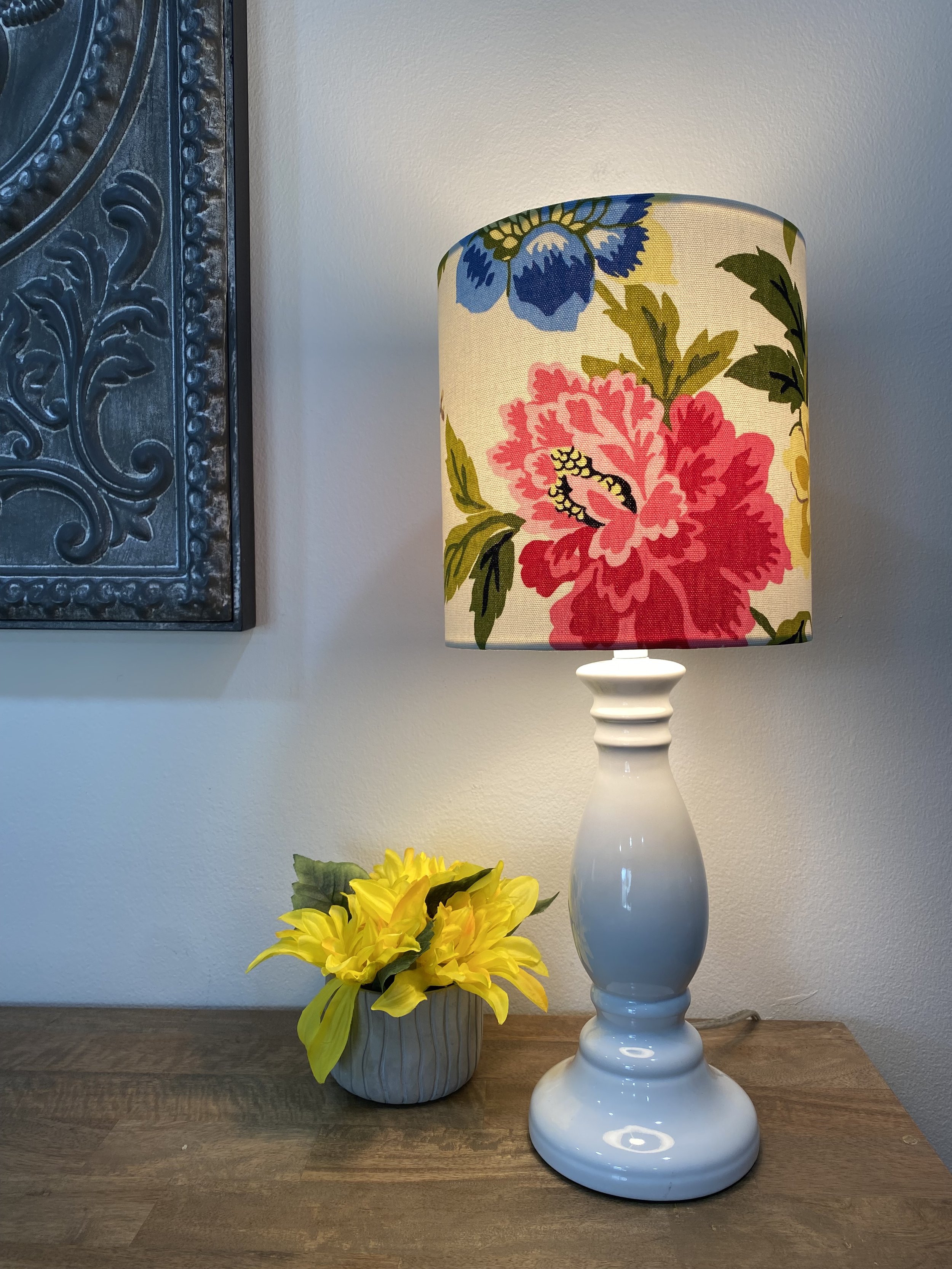 Table lamp with floral-patterned shade and white base, next to a small pot of yellow flowers on a wooden surface.