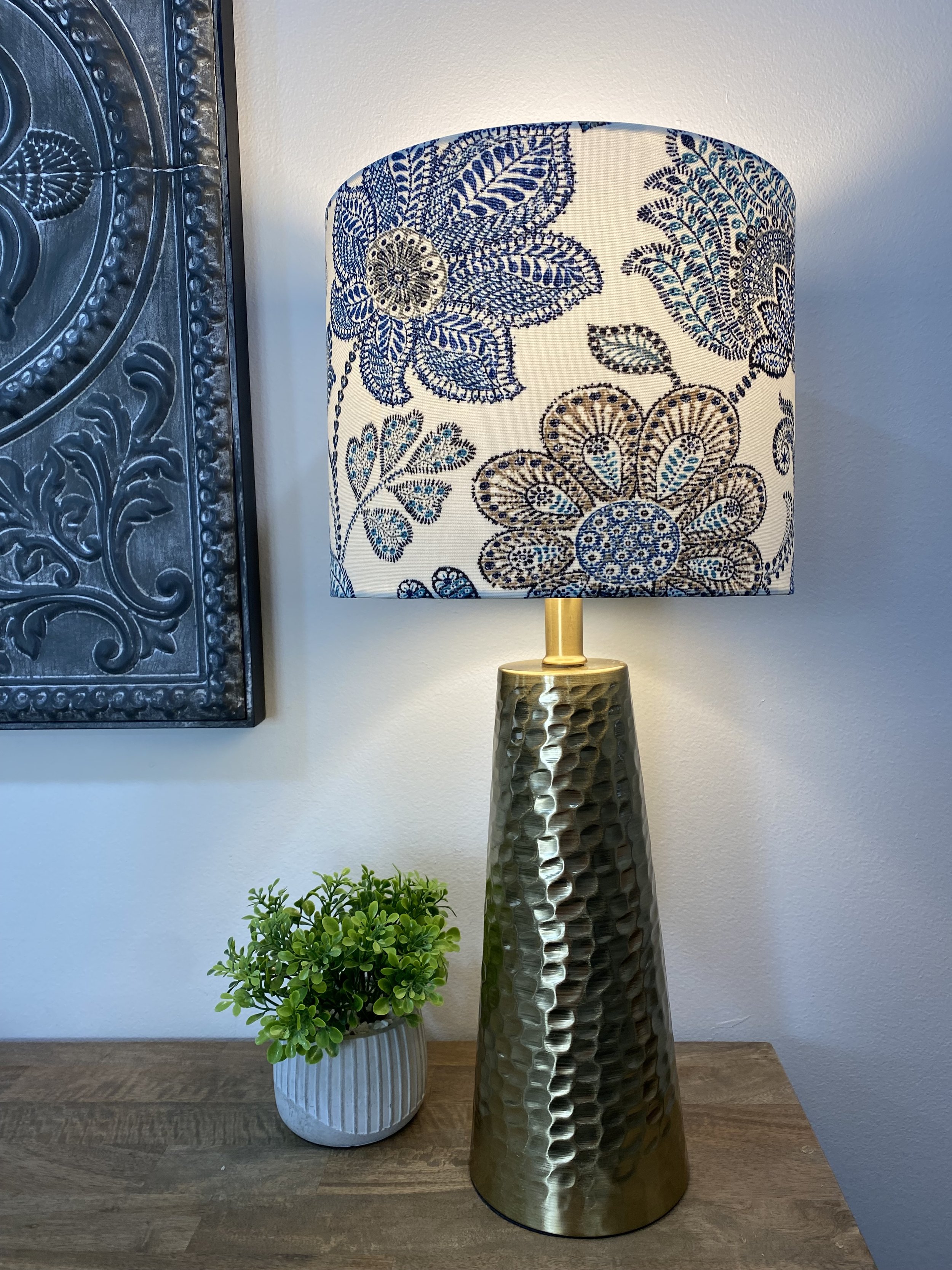 A brass lamp with a hammered texture and a floral-patterned lampshade sits on a wooden table. Next to it is a small potted green plant, and part of a decorative metal wall art piece is visible in the background.