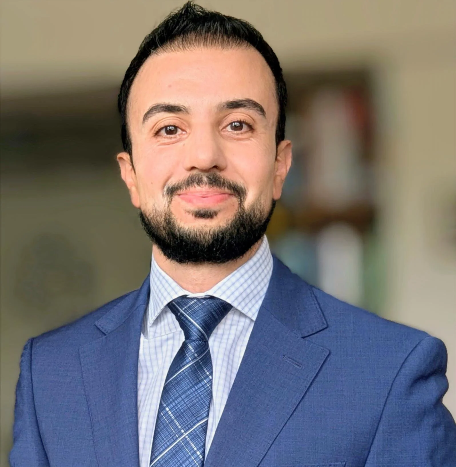 A man with dark hair and a beard, wearing a blue suit, a checked shirt, and a tie, smiling for a professional headshot.