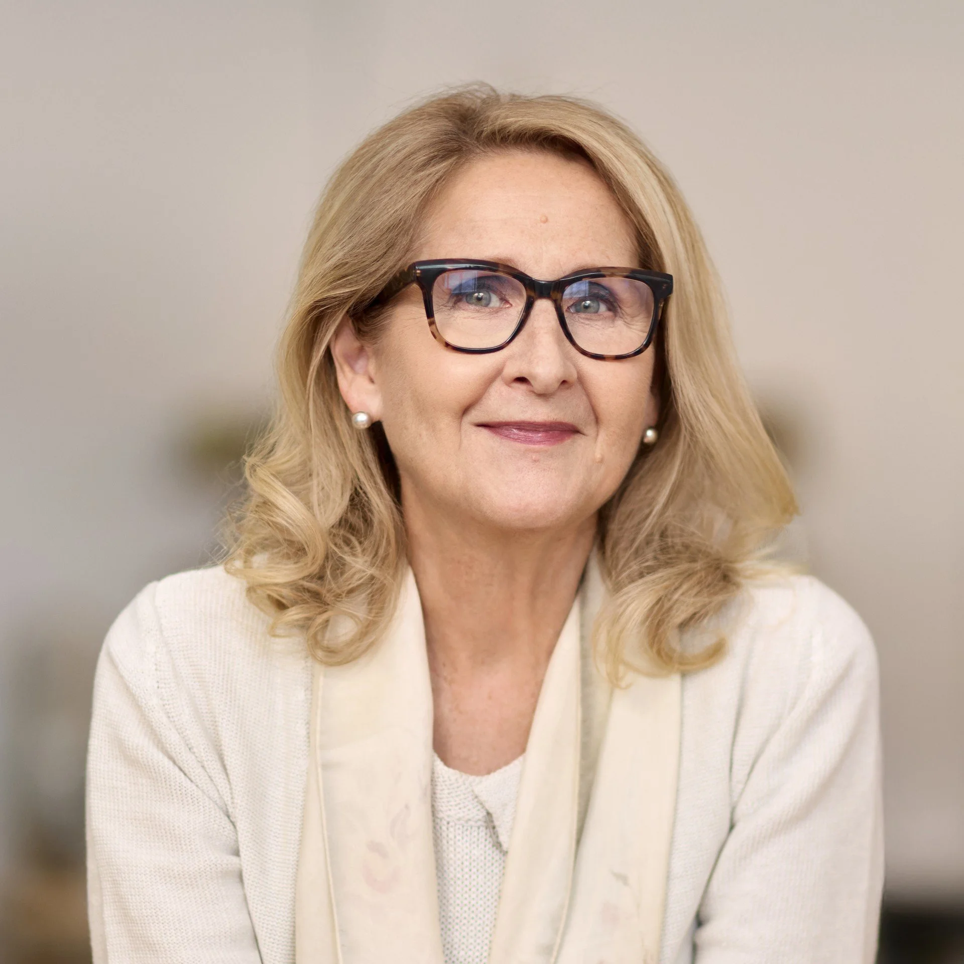 Une femme souriante, aux cheveux bouclés, portant des lunettes rouges, un collier de perles, une chemise blanche et un foulard coloré, dans un intérieur lumineux.