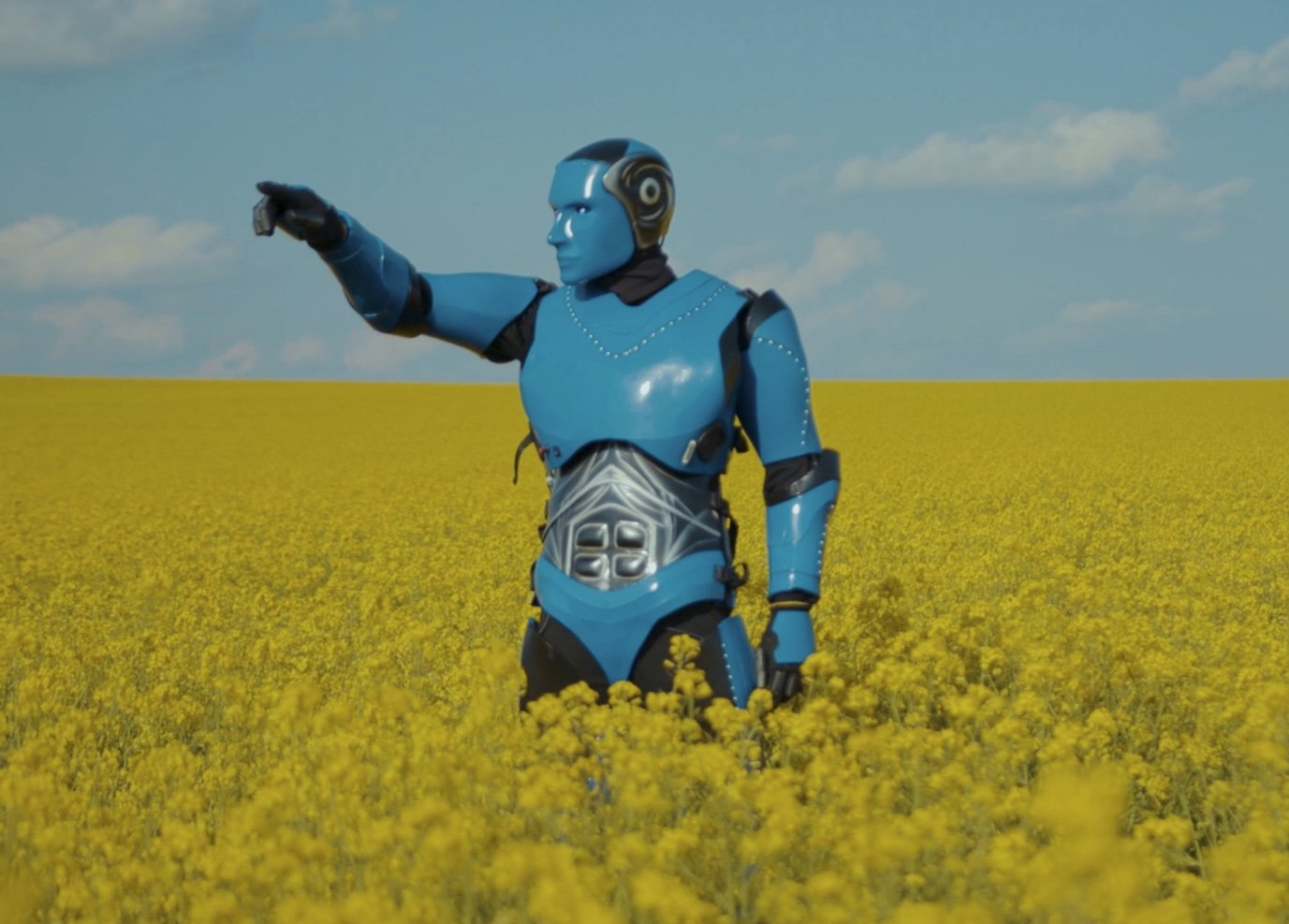 An MBI Energy Realty AI  humanoid robot with a blue exterior, standing in a yellow flower field under a partly cloudy sky, with one arm extended outward.