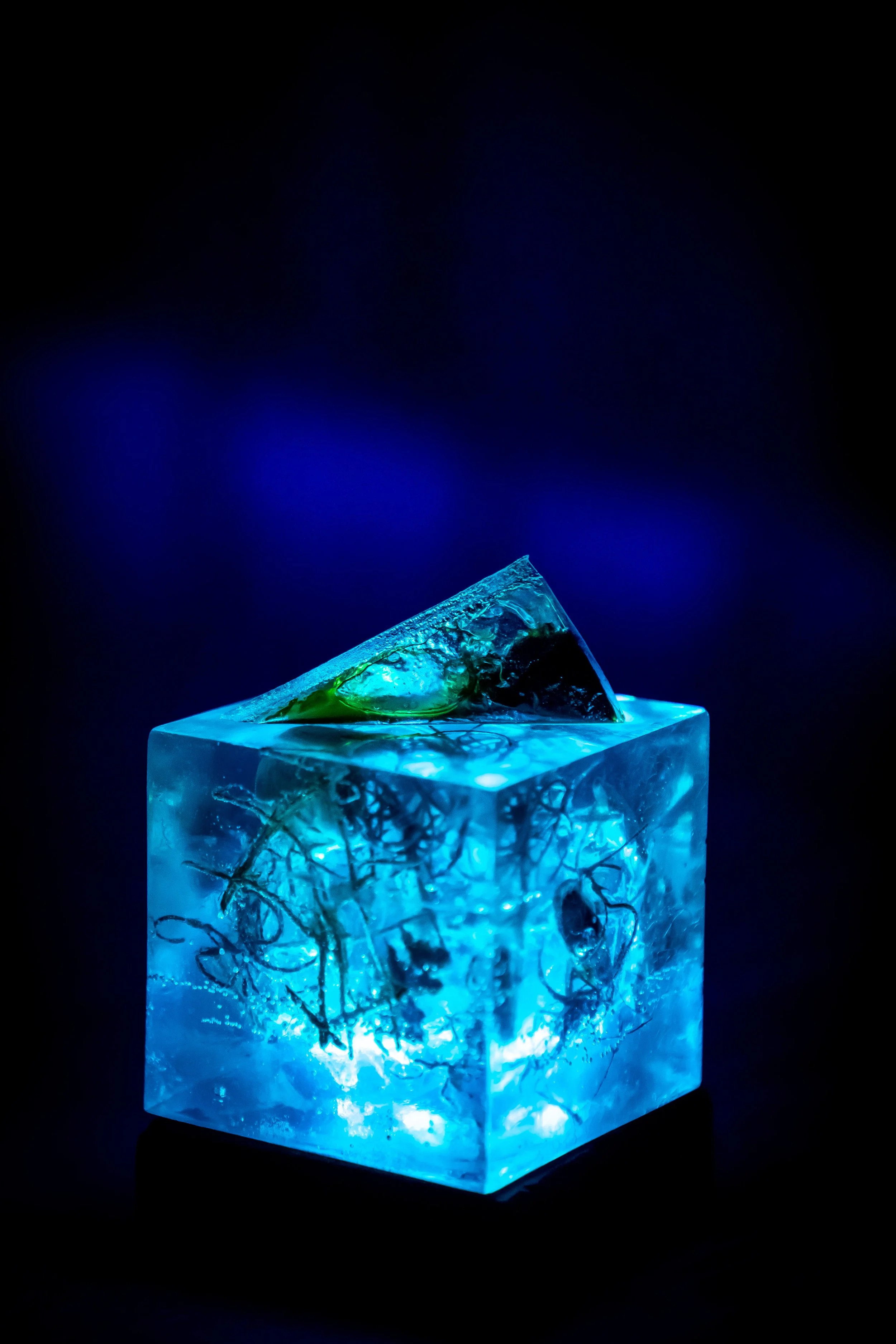 Close-up of a blue illuminated resin cube with internal structures and a reflective surface on top, against a dark blue background.