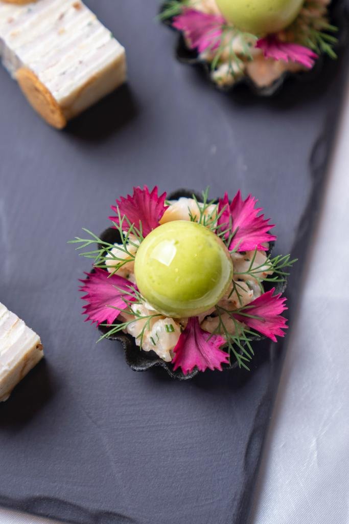 Gourmet appetizer with green mousse sphere, garnished with pink flower petals and herbs on a black slate plate.