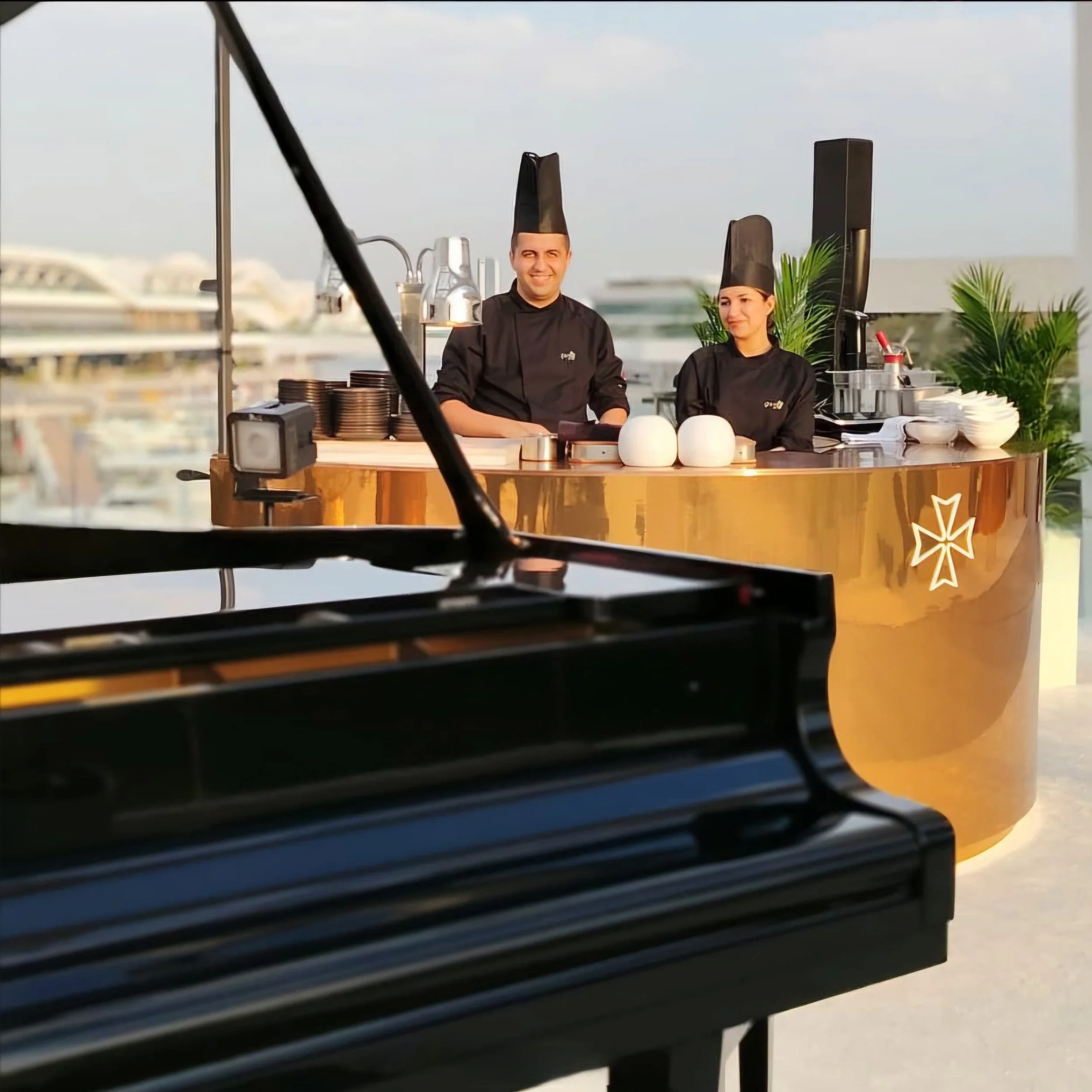 Chefs in black uniforms and hats stand behind a gold countertop with dishes, next to a black grand piano, outdoor setting with city view.