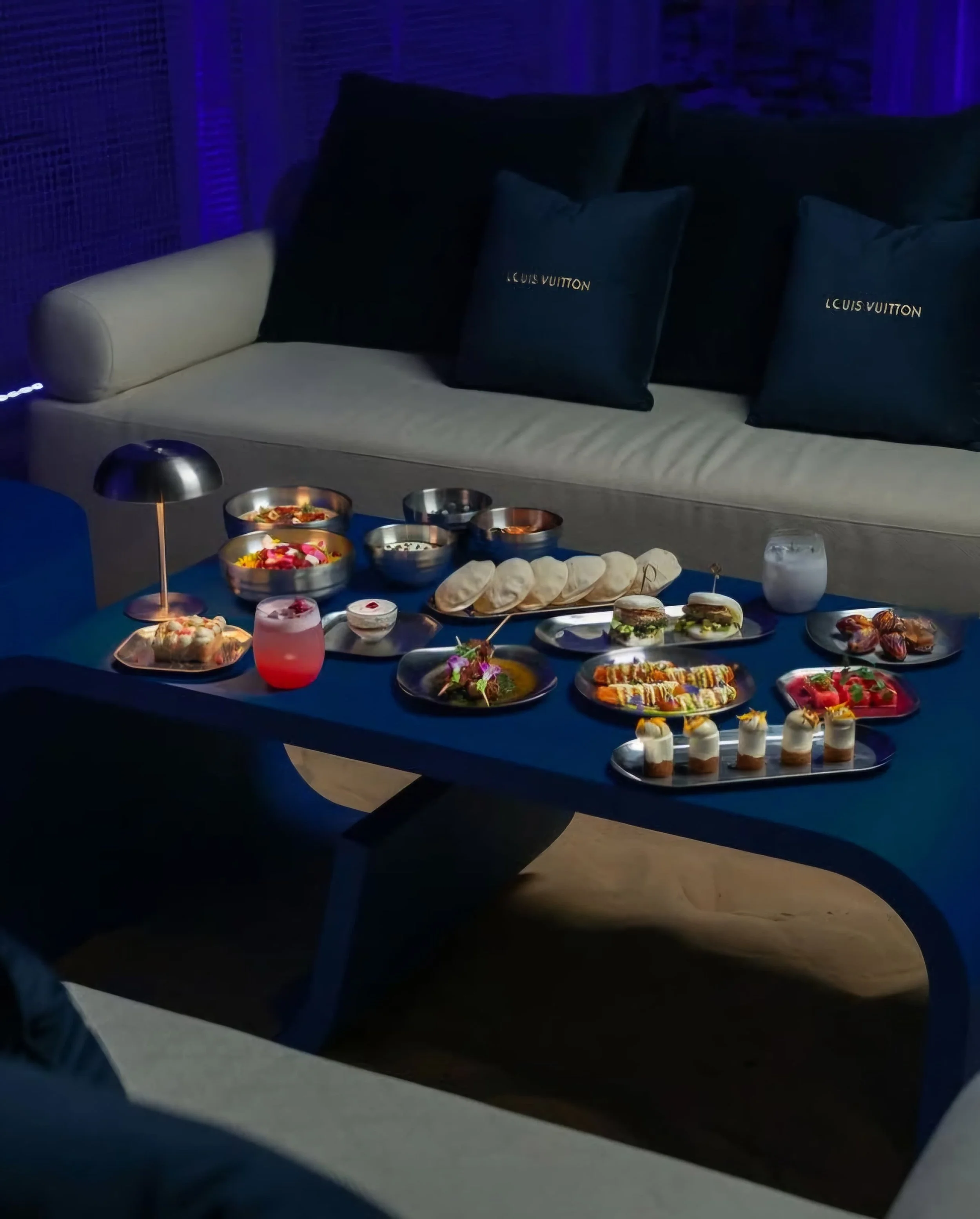 Elegant table setting with a variety of plated foods, drinks, and a small lamp, on a blue table in a sophisticated lounge with Louis Vuitton pillows.