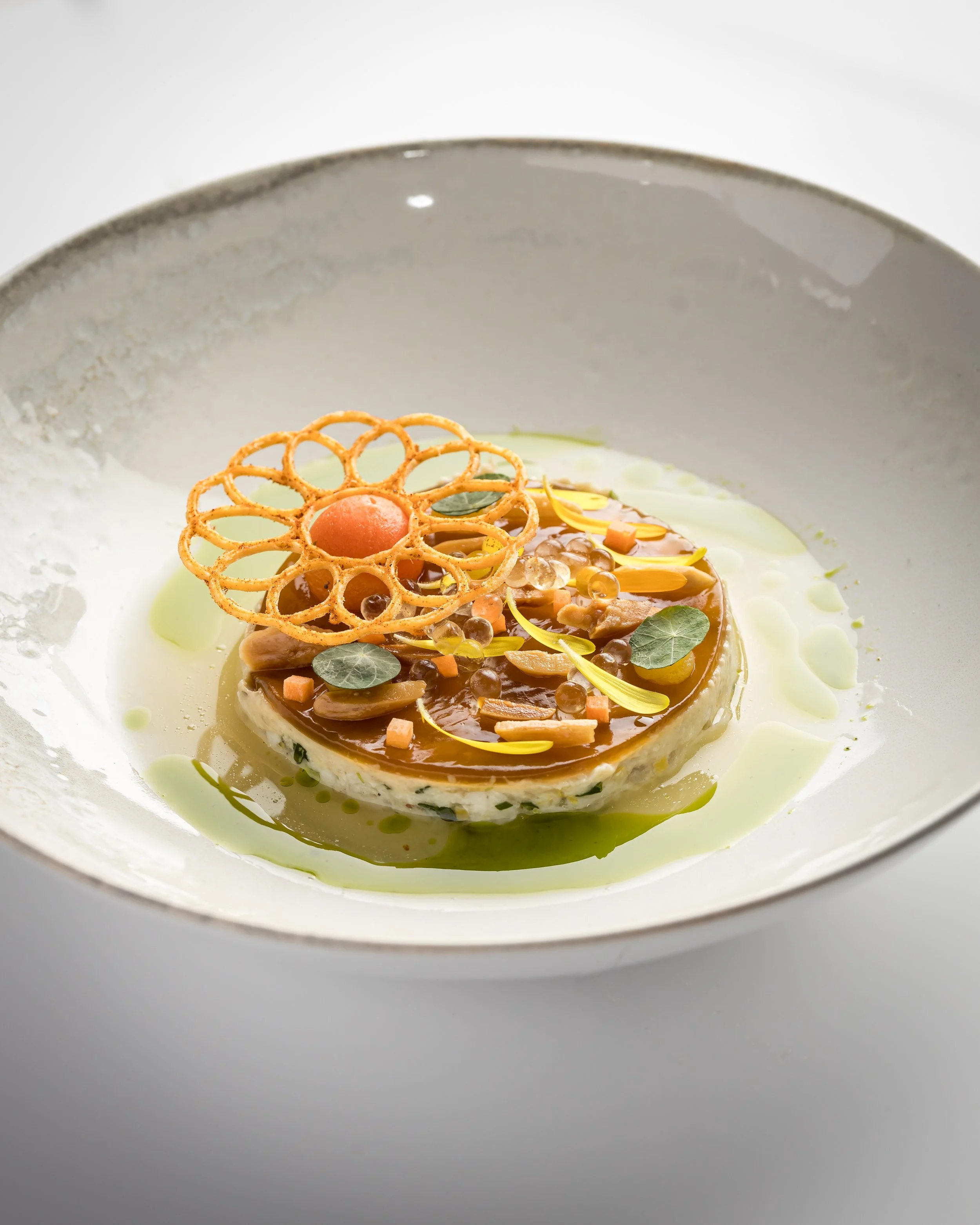 Gourmet dish with a round, creamy base topped with decorative elements, including a lattice pastry, edible flower petals, small herbs, and colorful garnishes, served in a white bowl with a green sauce.
