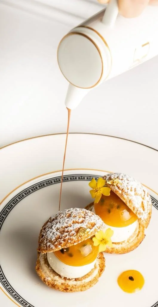 Gourmet dessert with choux pastries, cream, and fruit gel, garnished with yellow flowers and powdered sugar, with sauce being poured from a white pitcher.
