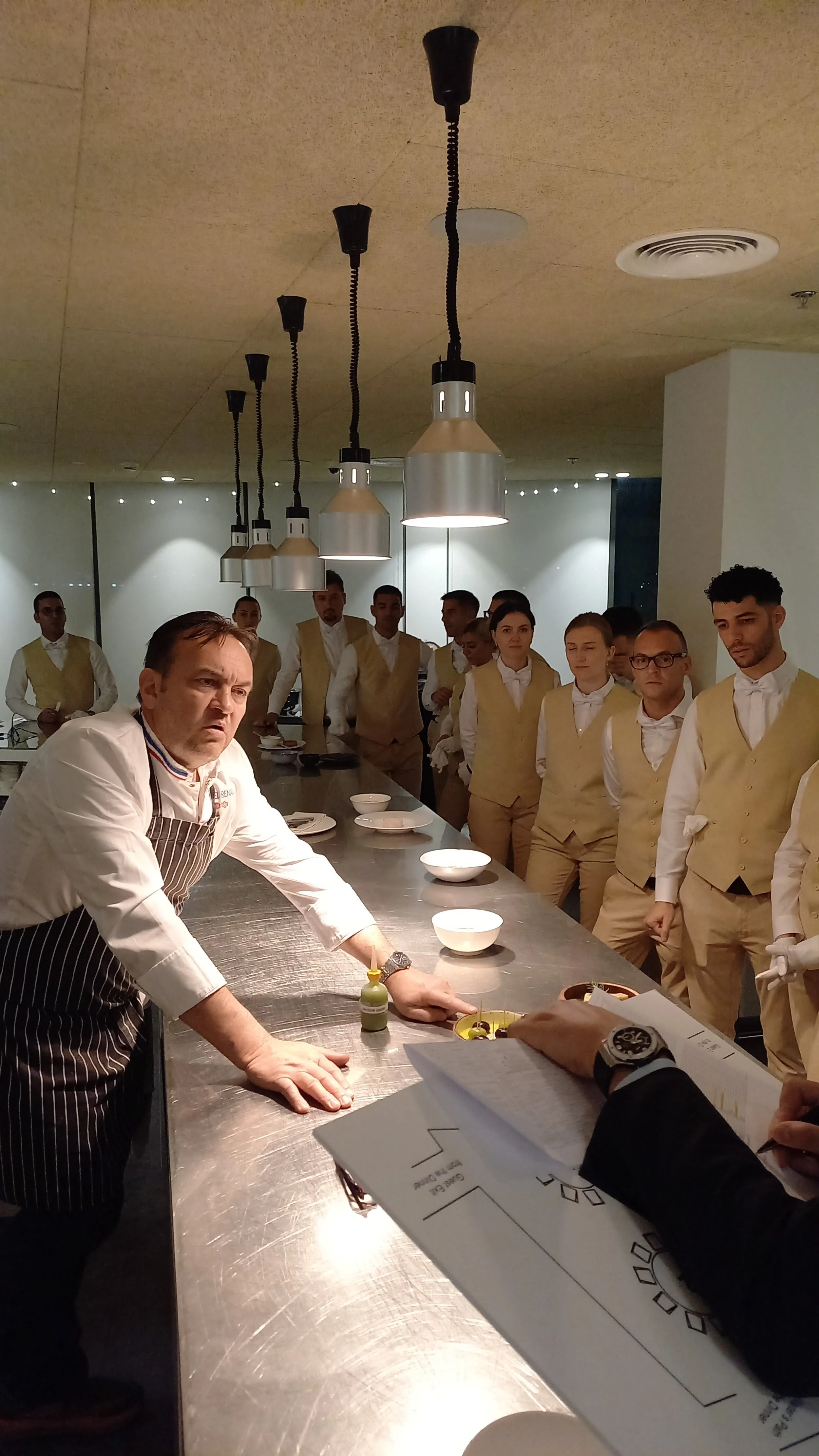 Chef instructing staff in a restaurant kitchen, kitchen layout on clipboard, wearing uniforms, standing around a stainless steel table.