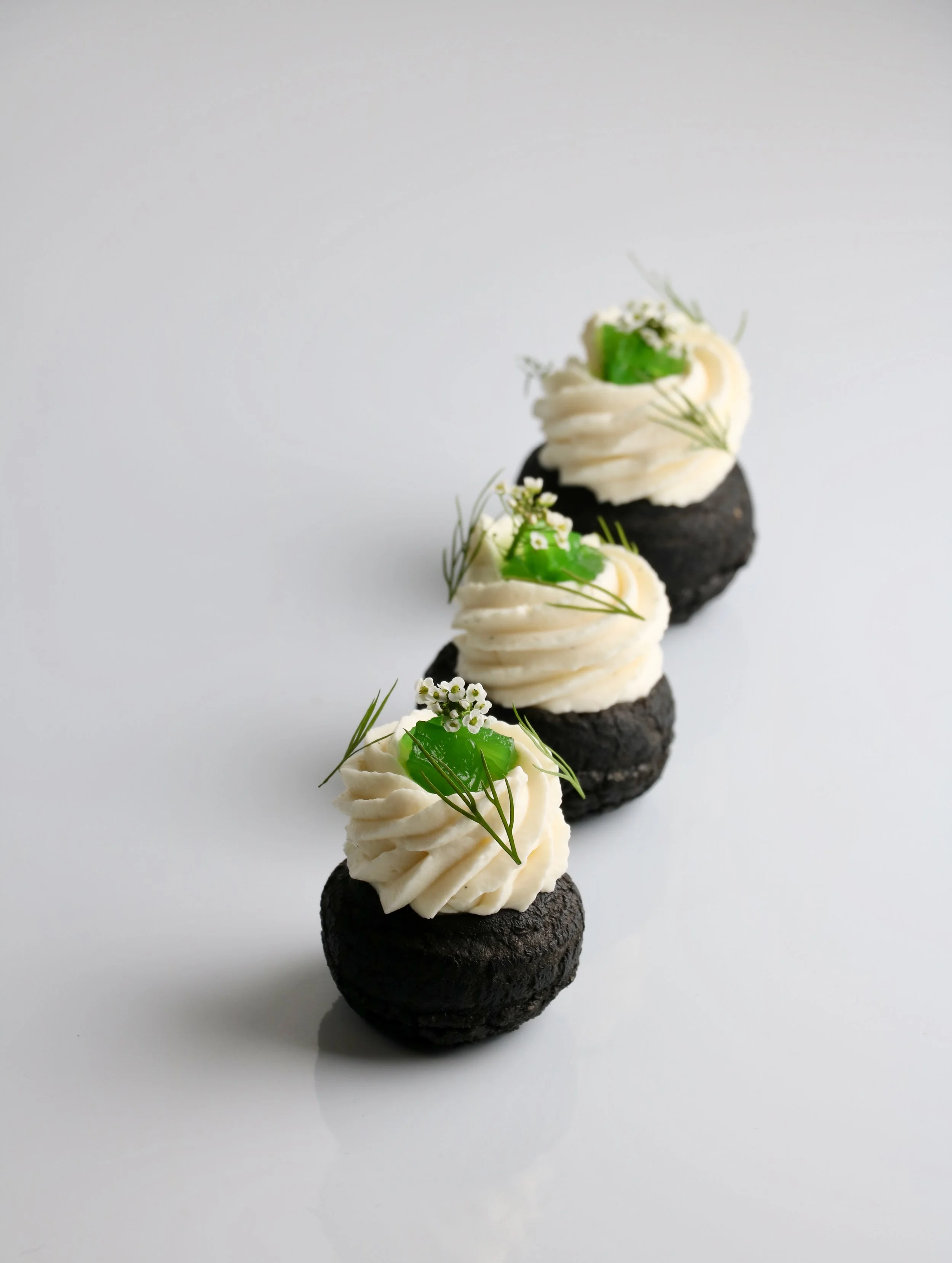 Gourmet charcoal biscuits topped with white cream, green jelly, and garnished with herbs and small flowers, arranged on a reflective white surface.