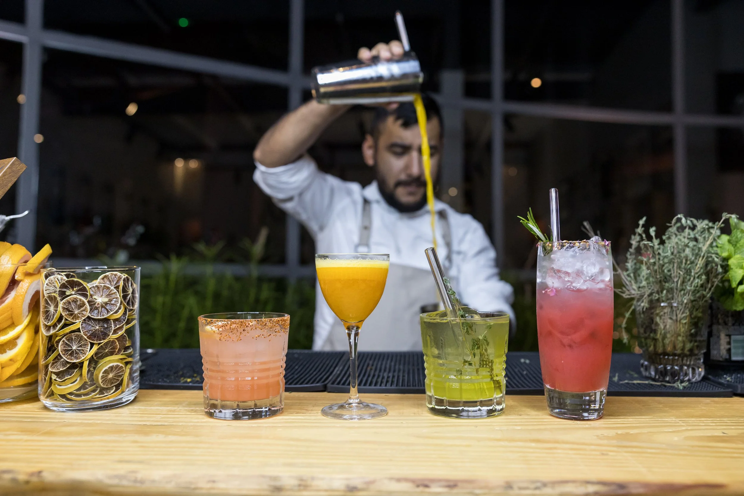Bartender pouring a drink surrounded by colorful cocktails on a wooden bar with garnish jars.