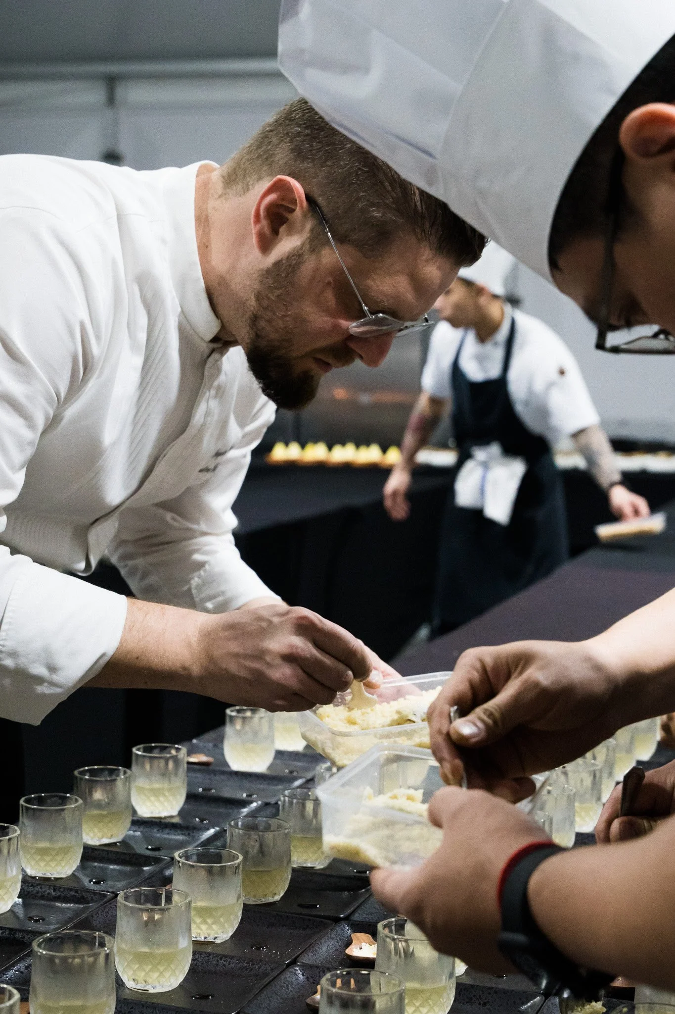 Chefs preparing food in a professional kitchen, arranging ingredients in glasses on a counter.