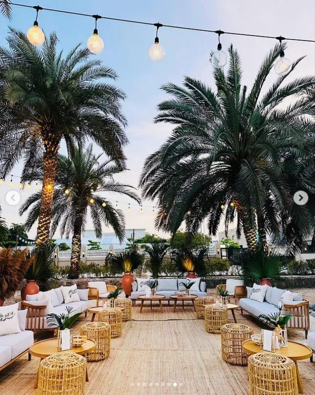 Outdoor lounge area with sofas, wicker tables, and palm trees under string lights at sunset.