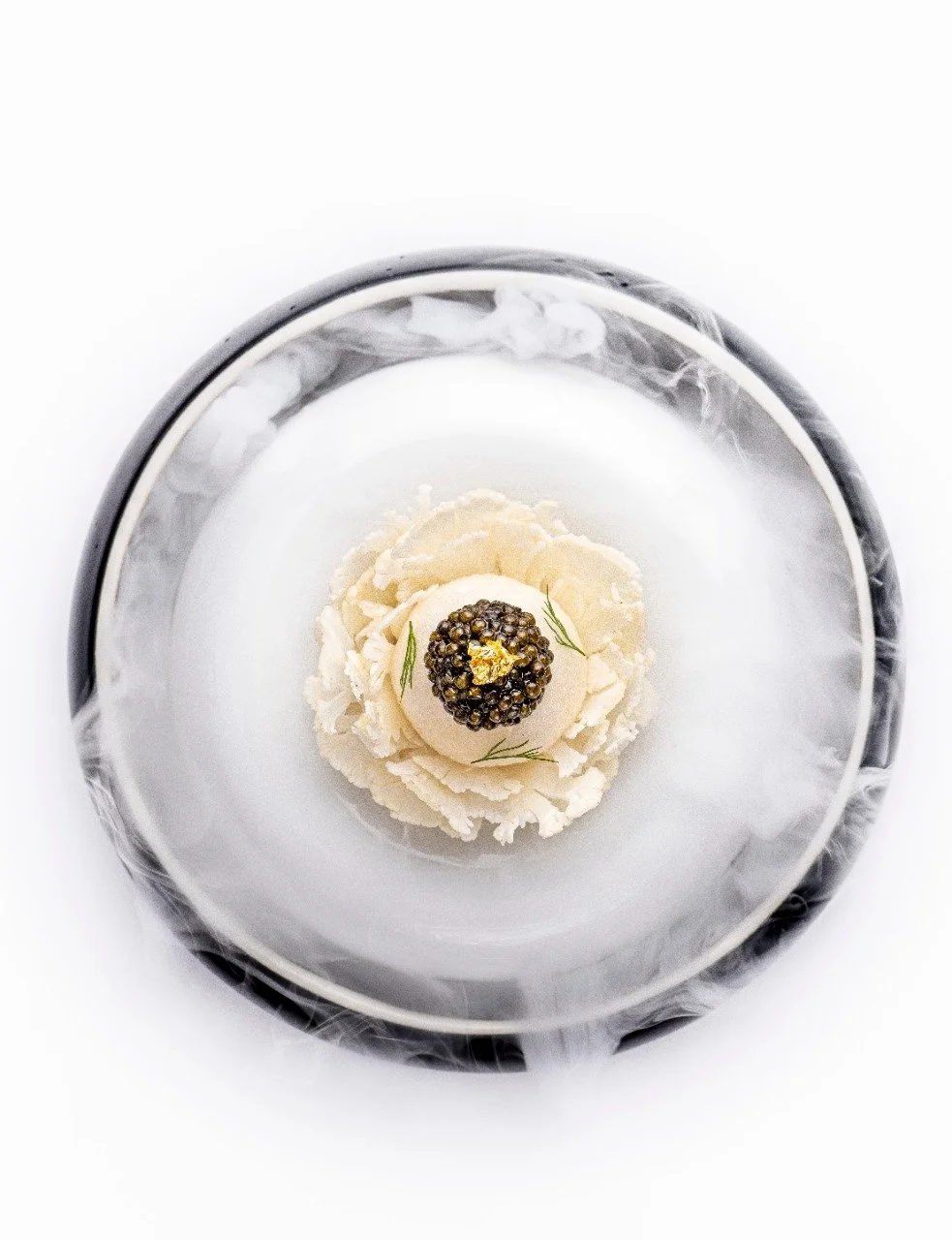 Elegant dish featuring caviar on cream, garnished with gold leaf and herbs, surrounded by decorative smoke on a black and white plate.