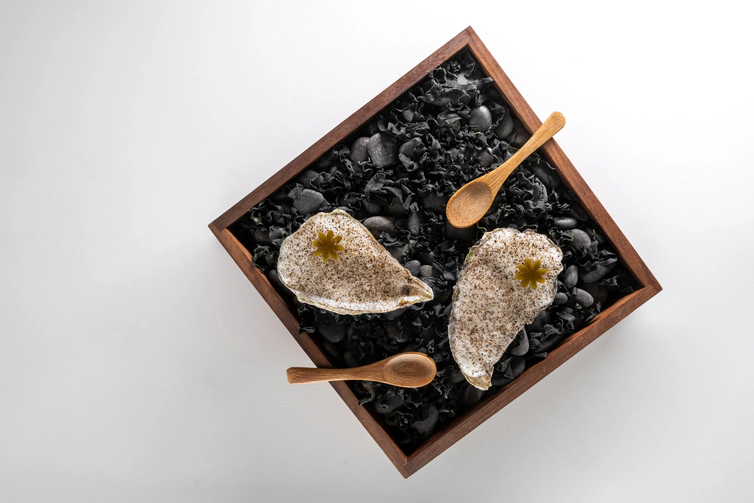 Two season oysters with garnish, displayed in a wooden box filled with black stones and seaweed, wooden spoons on the side.