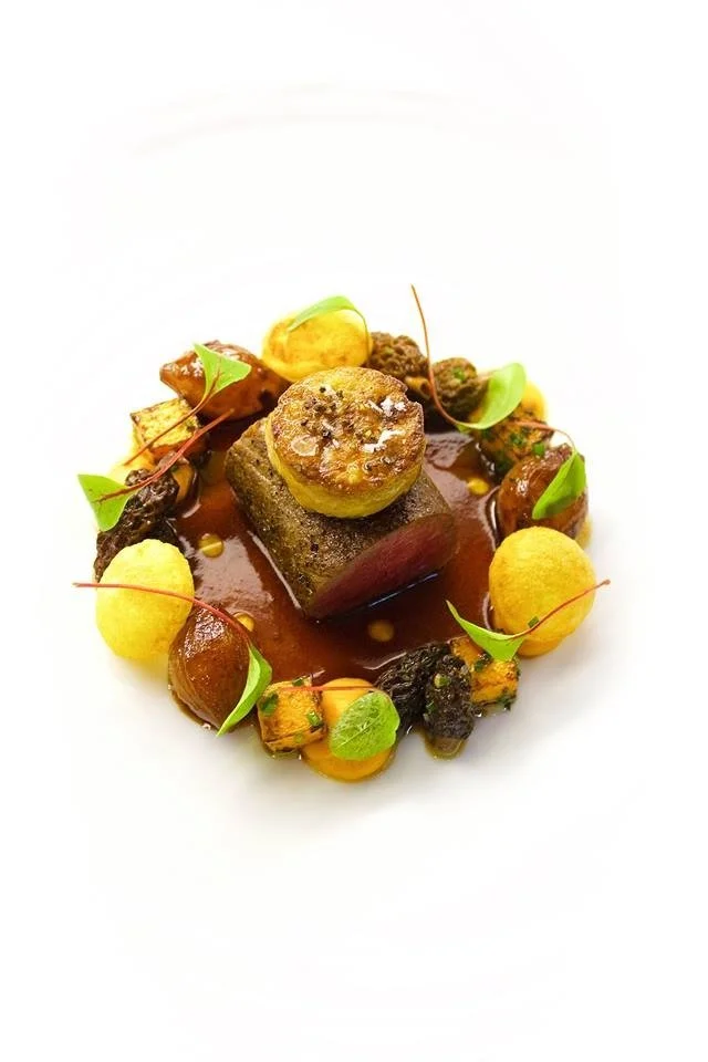 Gourmet dish with seared meat, sauce, vegetables, microgreens, and garnishes on a white plate.