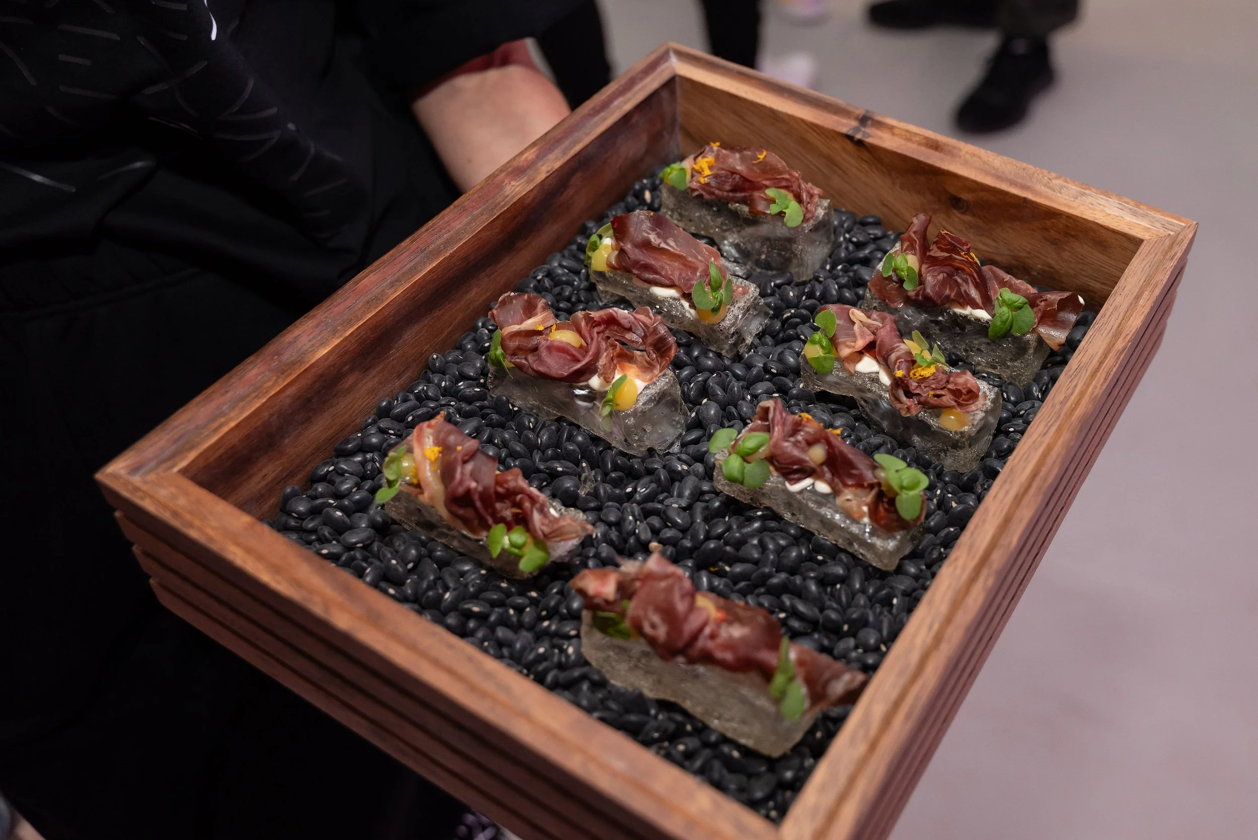Wooden tray with gourmet appetizers on ice blocks, black beans, garnished with herbs.