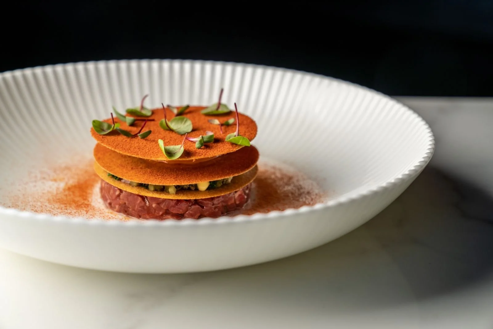 Gourmet dish in a white bowl with layers of crispy discs, microgreens, and a meat tartare base on a marble surface.