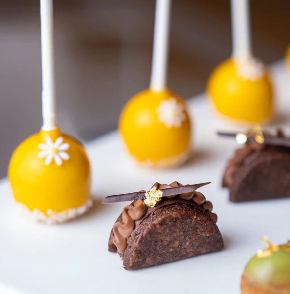 Close-up of yellow cake pops with daisy decorations and mini chocolate desserts on a white surface.